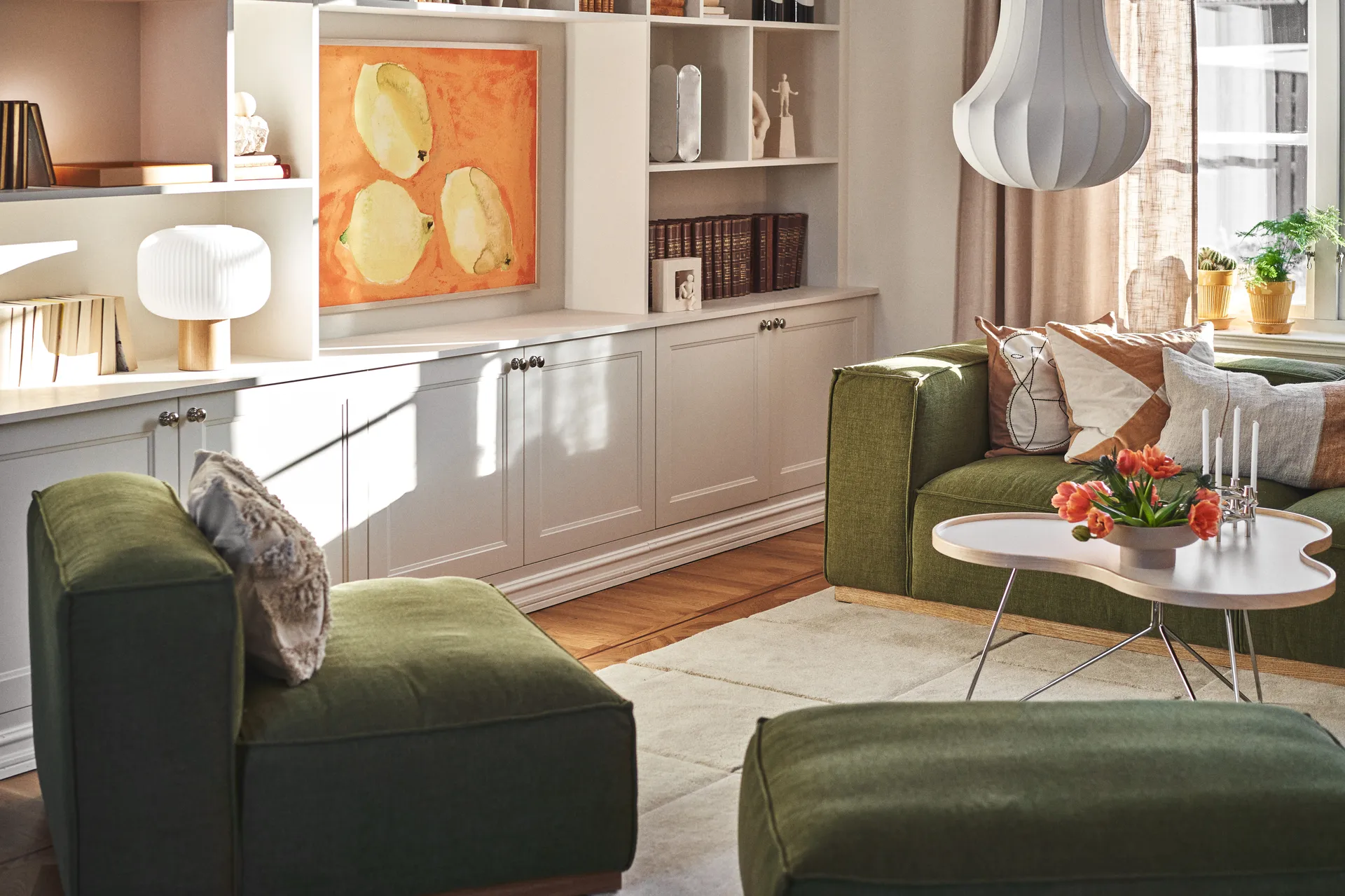 Salon ensoleillé avec canapés verts, une table basse blanche avec tulipes oranges et bougies, et des étagères blanches avec de l'art.