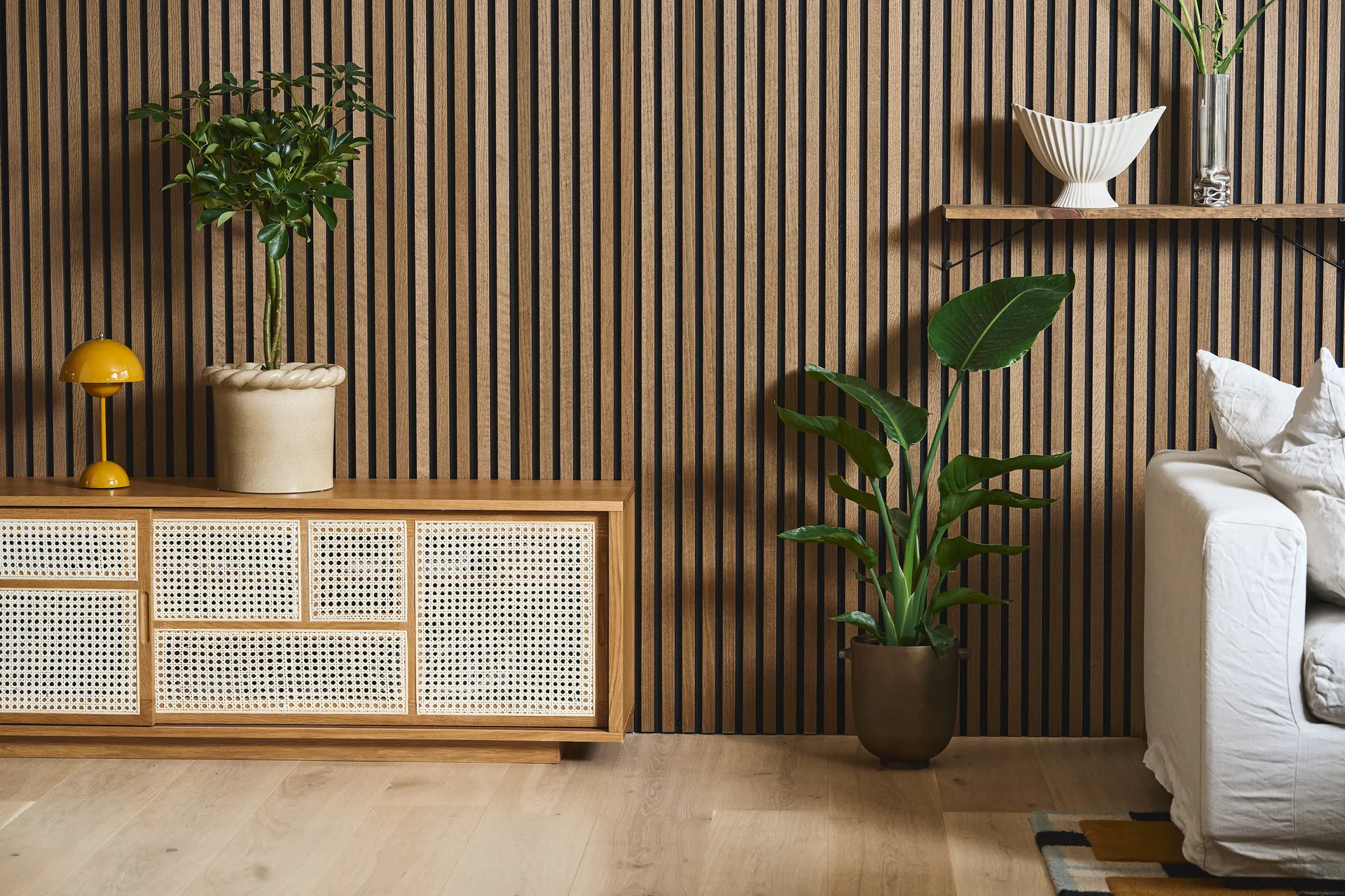 Salon moderne avec mur à lattes de bois, meuble en rotin, lampe jaune, plantes en pot et canapé blanc.