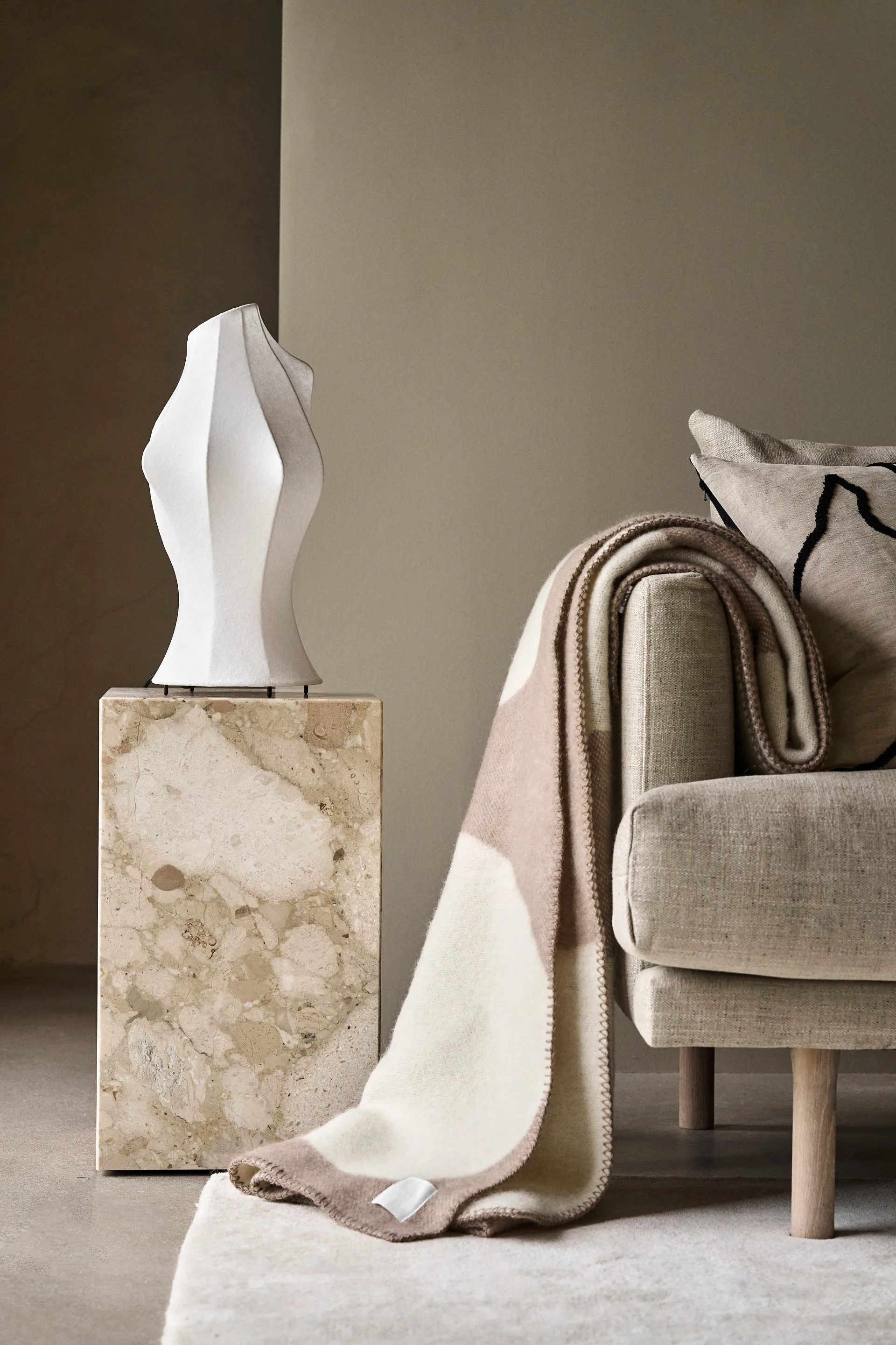 Lampe sculpturale blanche sur piédestal en marbre, à côté d'un canapé beige clair avec un plaid crème et marron et un coussin.