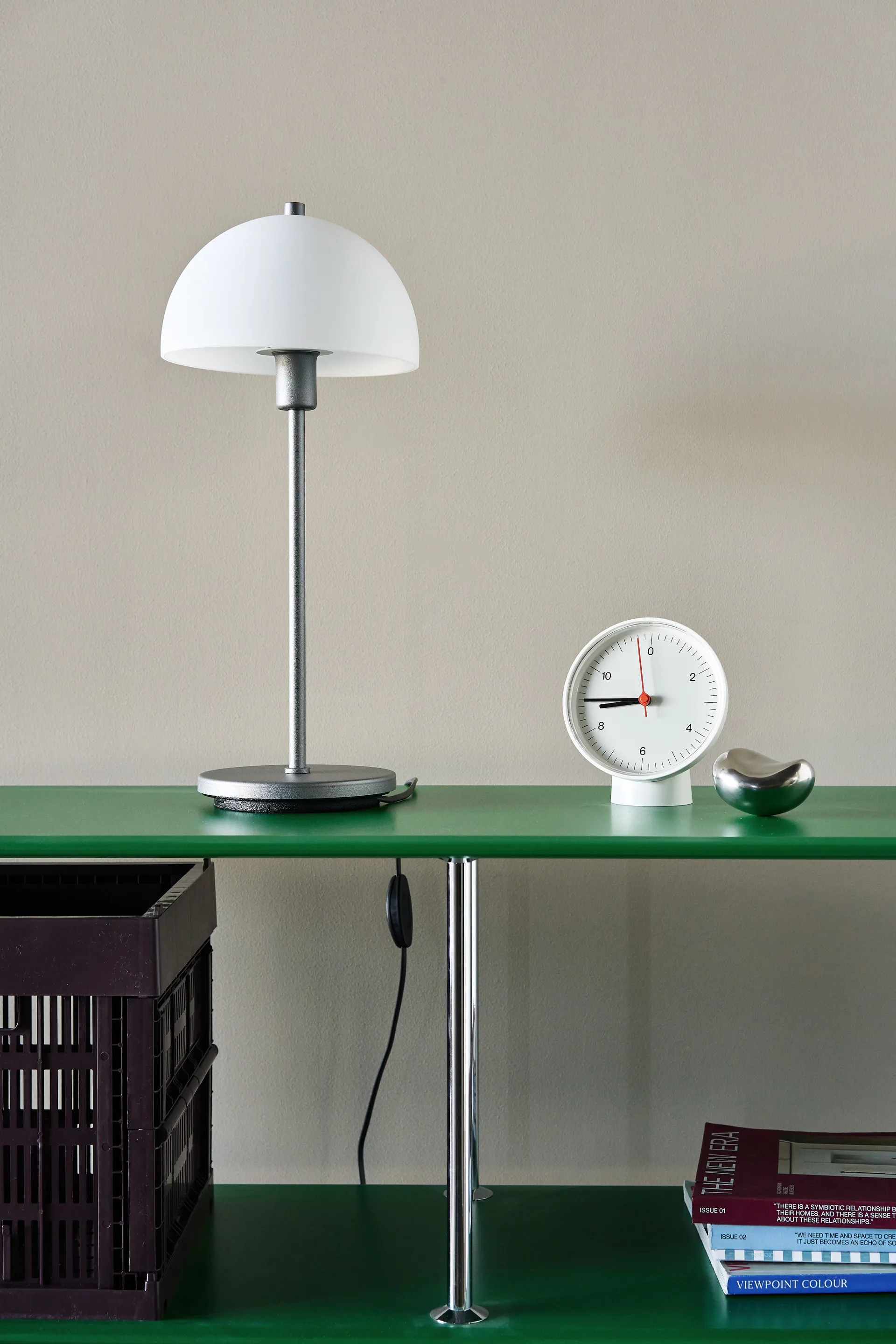 Étagère verte minimaliste avec lampe blanche en dôme, horloge ronde blanche et galet argenté. Caisse sombre et magazines en dessous.