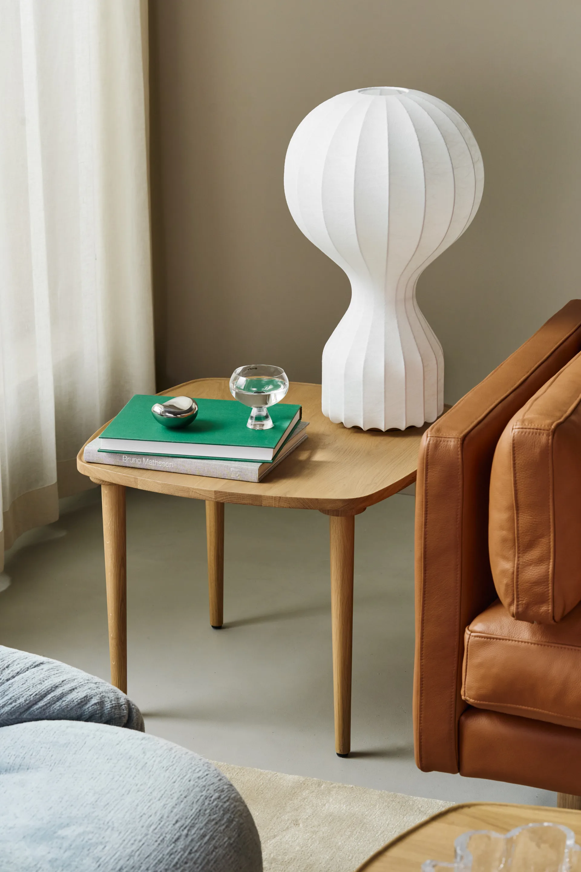 Table d'appoint en bois clair avec lampe blanche nervurée, livres verts, verre d'eau et objet argenté, à côté d'un fauteuil en cuir marron.