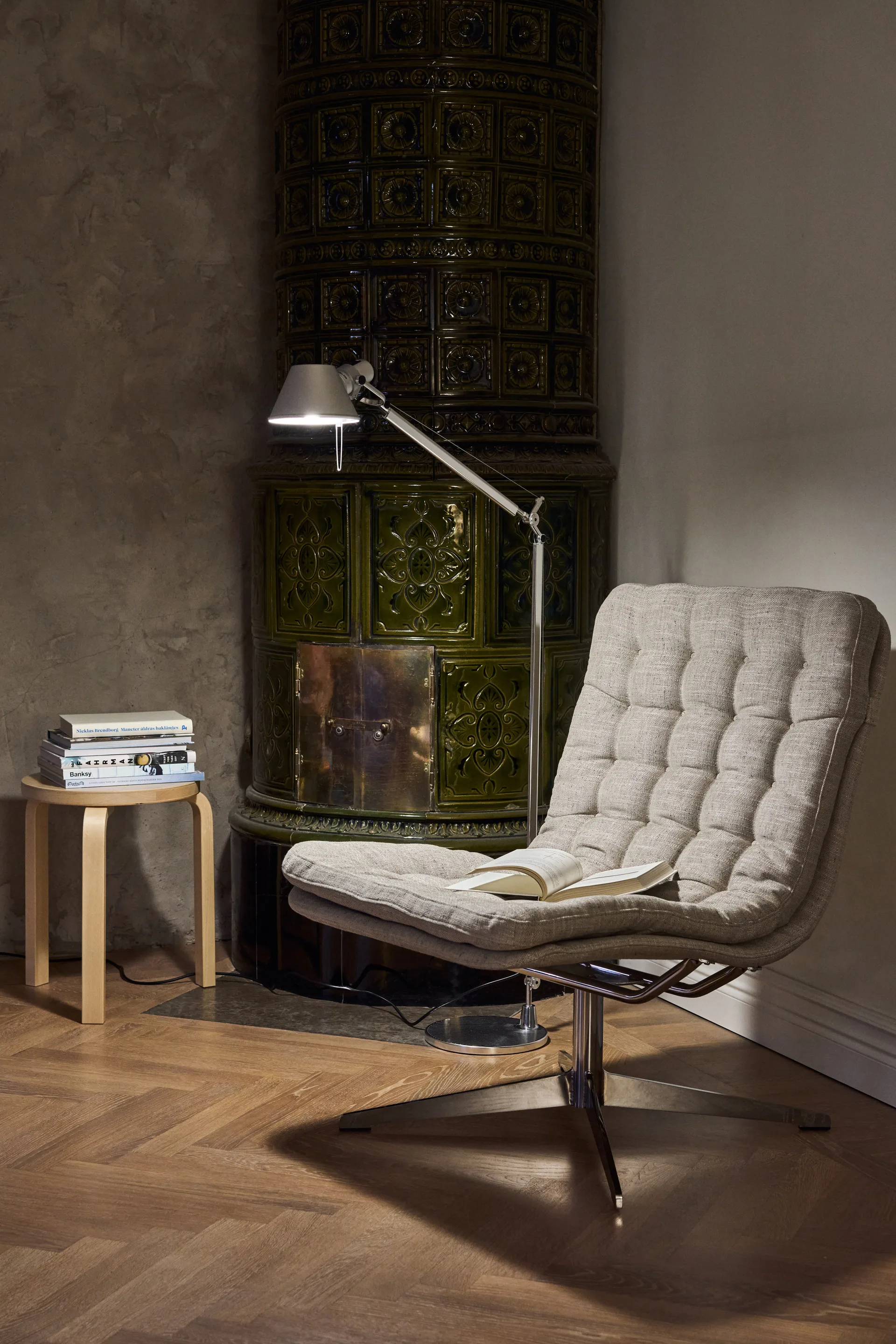 Fauteuil pivotant gris et lampadaire argenté à côté d'un poêle en faïence verte.