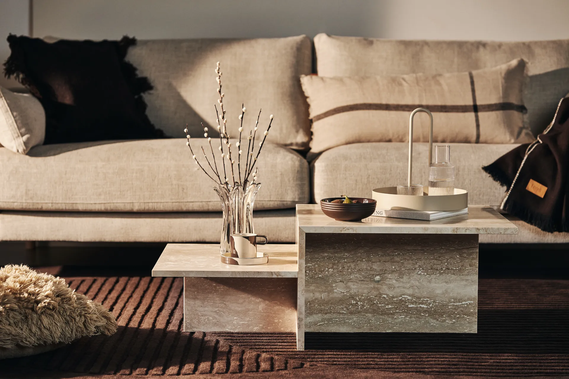Table basse à niveaux en pierre beige devant un canapé neutre avec des accessoires décoratifs.