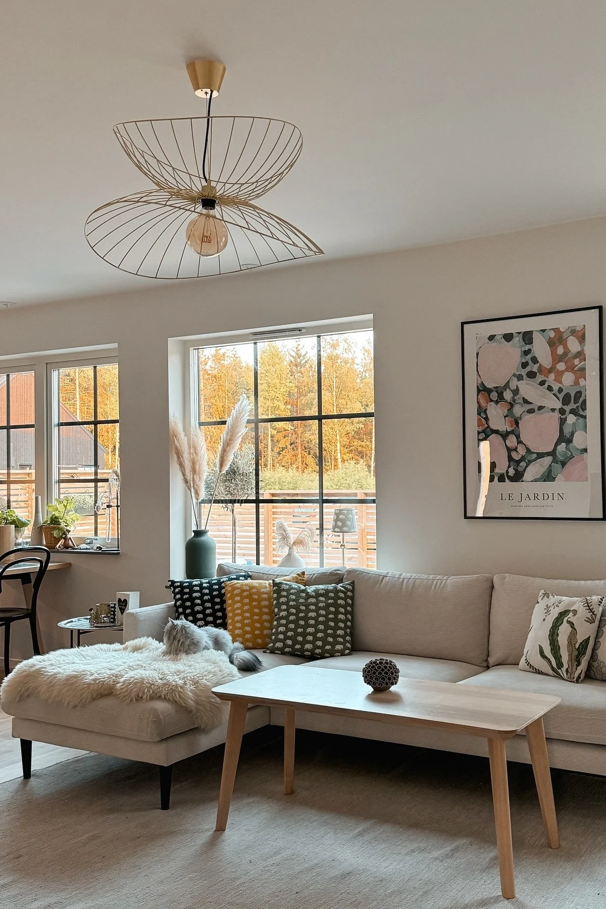 Dans un salon scandinave joyeux avec canapé, table basse et grandes fenêtres, est suspendu le plafonnier Ray de Globen Lighting.