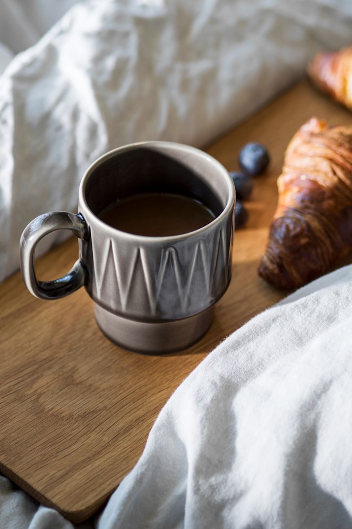 La tasse à espresso Coffee & More grise repose sur une planche en bois posée sur un lit, avec un croissant à côté.