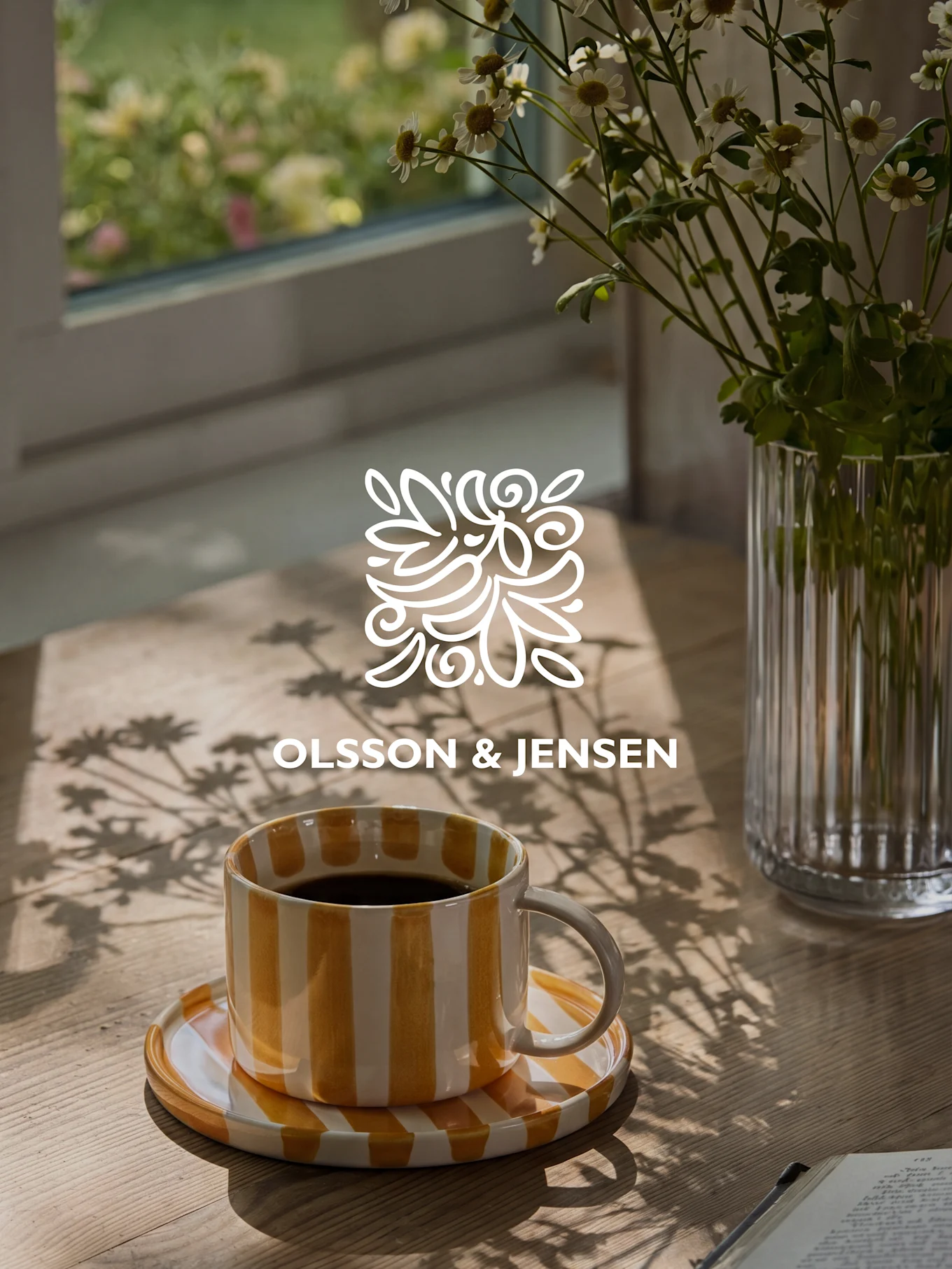 Tasse à café rayée jaune et blanche sur une table en bois avec un vase de fleurs blanches et des ombres ensoleillées, logo Olsson & Jensen.