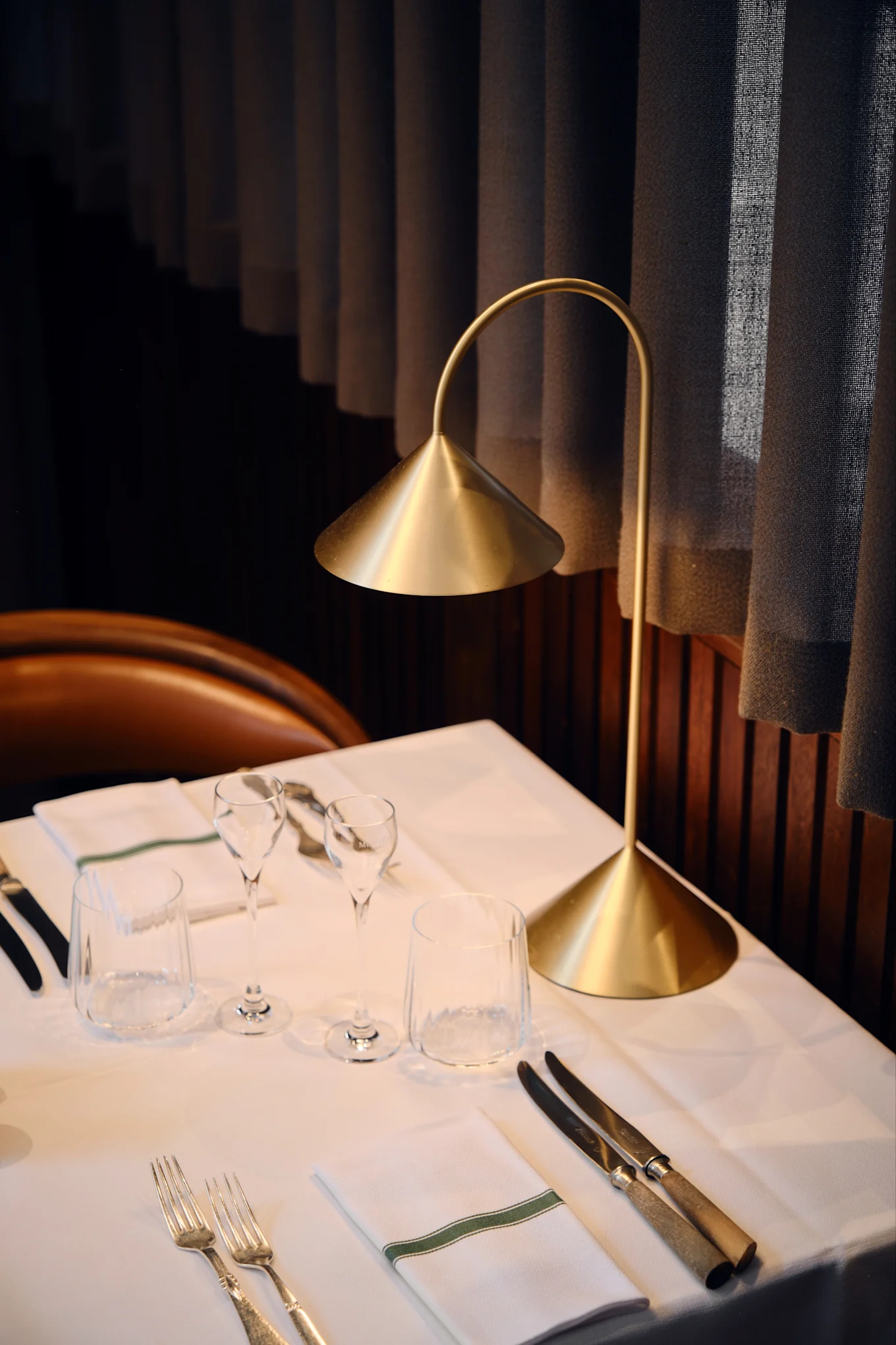 Une lampe de table dorée moderne illumine une table de restaurant dressée avec du linge blanc, des verres, des couverts et une serviette à rayure verte.