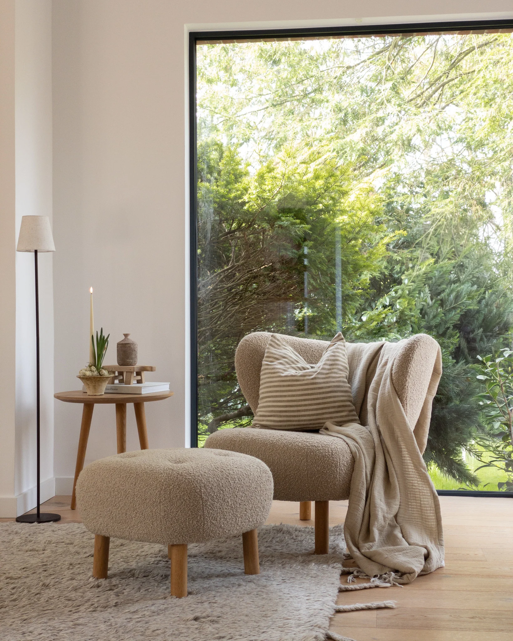 Coin douillet : fauteuil marron clair, pouf, table d'appoint avec bougie, grande fenêtre, arbres verts.