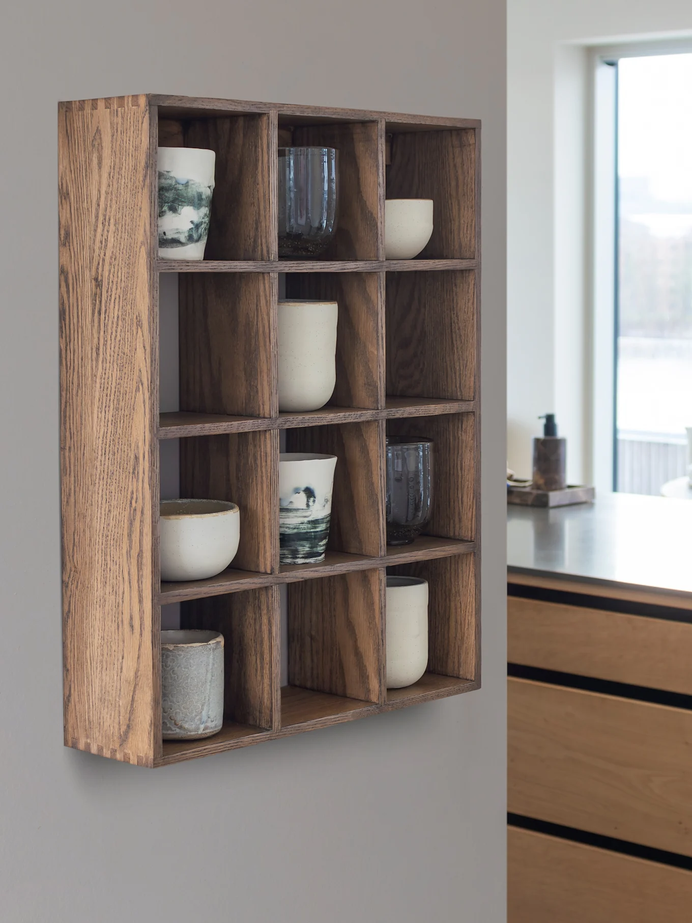Étagère murale en bois avec 12 compartiments contenant des tasses et des mugs en céramique. Une cuisine moderne et une fenêtre sont en arrière-plan.