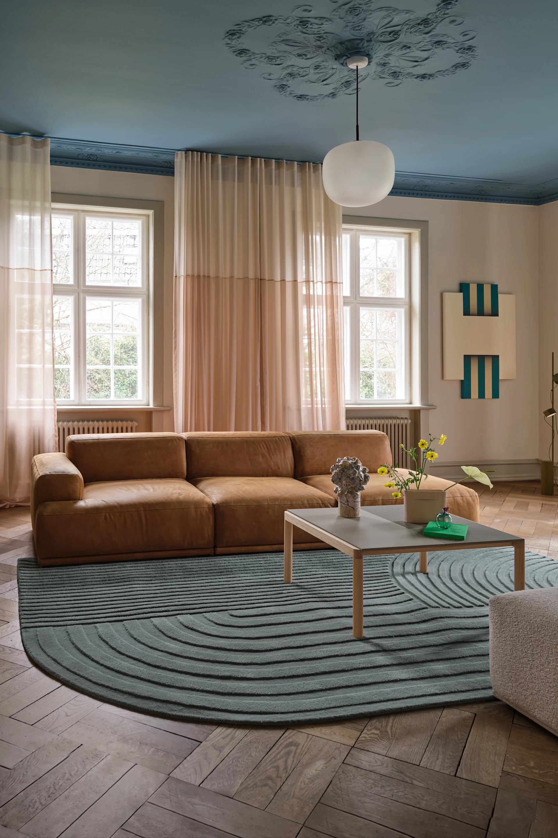 Salon moderne : canapé en cuir marron, tapis à motifs turquoise, plafond bleu clair avec un lustre, grandes fenêtres.