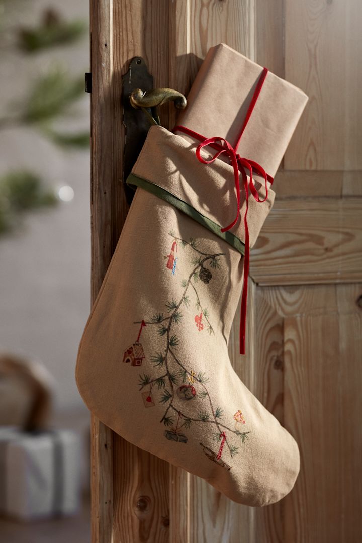 Les décorations de Noël pour 2025 s'inspirent de la nature, comme cette chaussette de Kähler que vous voyez ici suspendue à une porte en bois devant un sapin de Noël et des cadeaux emballés. 