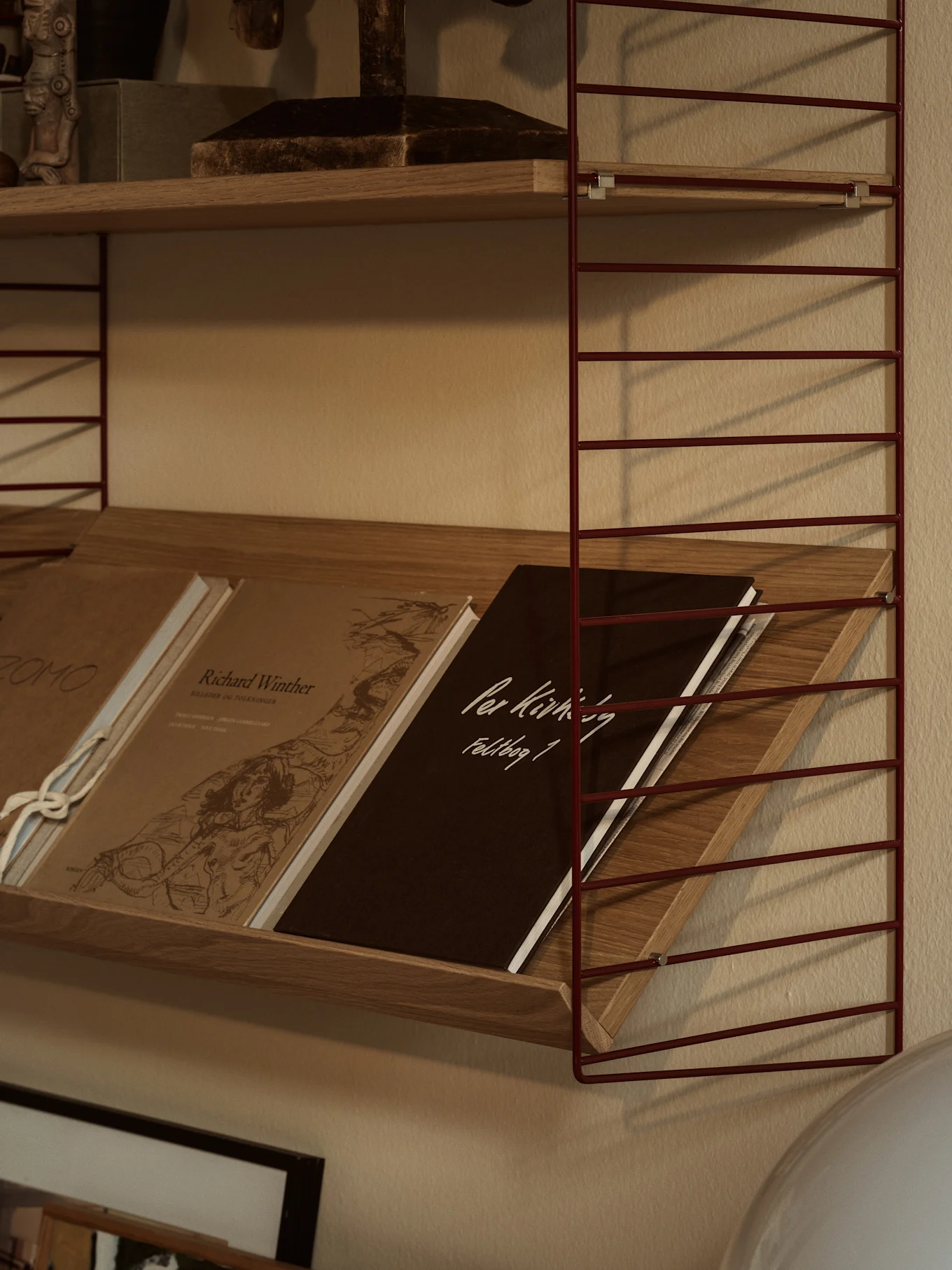 Une étagère murale à fils métalliques rouges avec des étagères en bois. L'étagère inférieure contient des livres inclinés, dont un marron foncé avec 'Per Kirkeby Feldbog 1'.