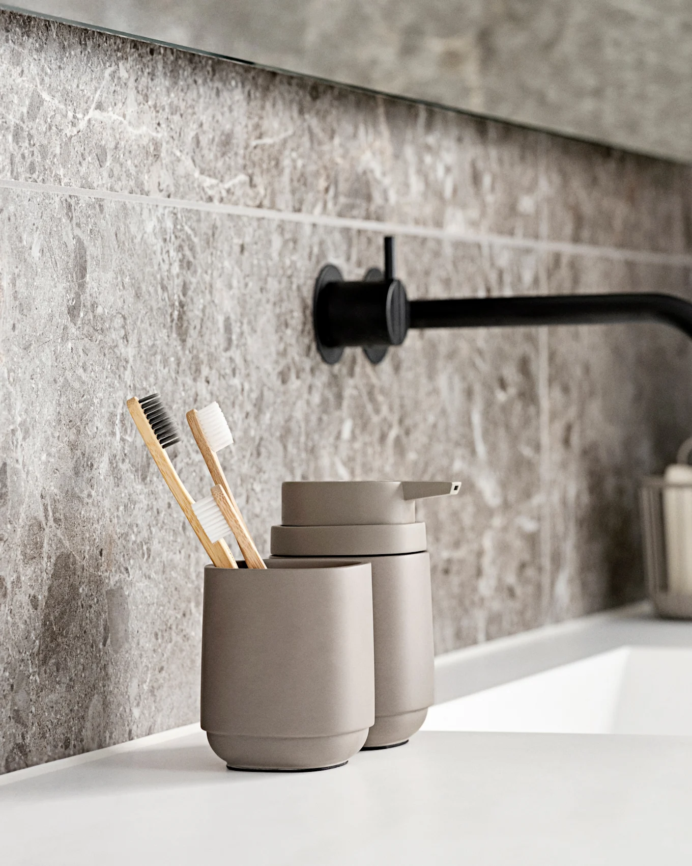 Salle de bain moderne avec lavabo blanc, robinet mural noir, carreaux aspect marbre gris et brosses à dents écologiques.