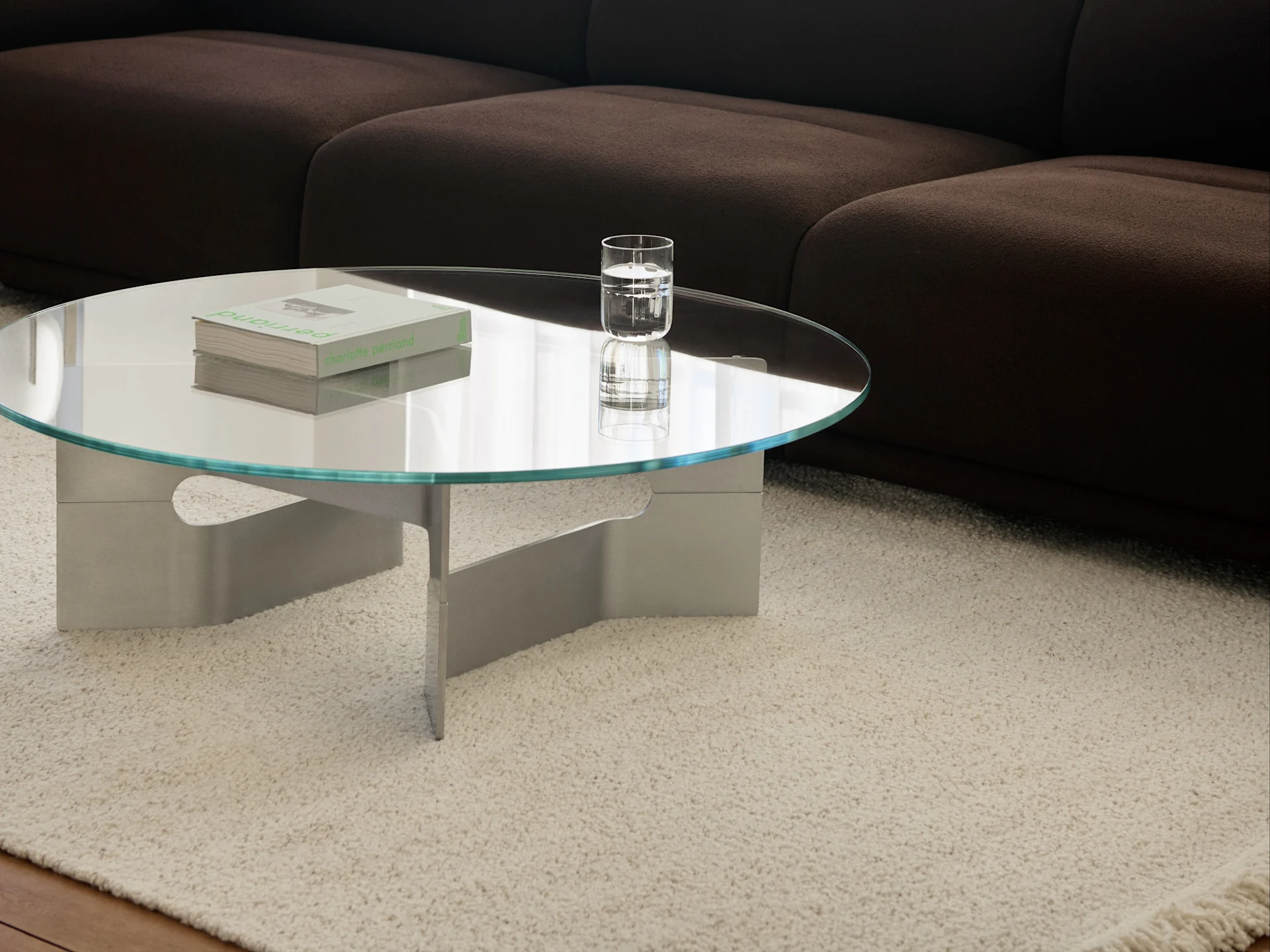 Table basse ronde en verre avec des livres et un verre d'eau sur un tapis shaggy crème devant un canapé marron foncé.