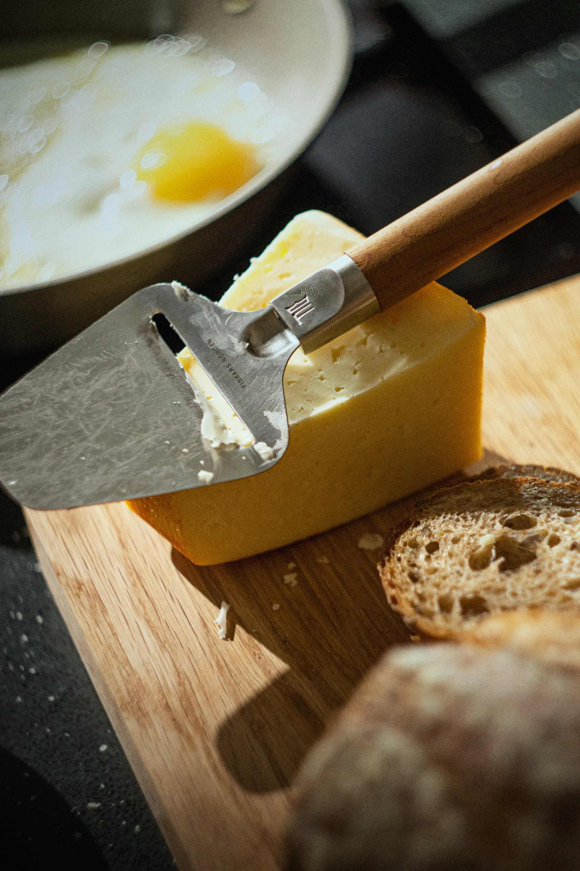 Une trancheuse à fromage repose sur un bloc de fromage jaune sur une planche en bois, avec des tranches de pain et un œuf au plat dans une poêle en arrière-plan.