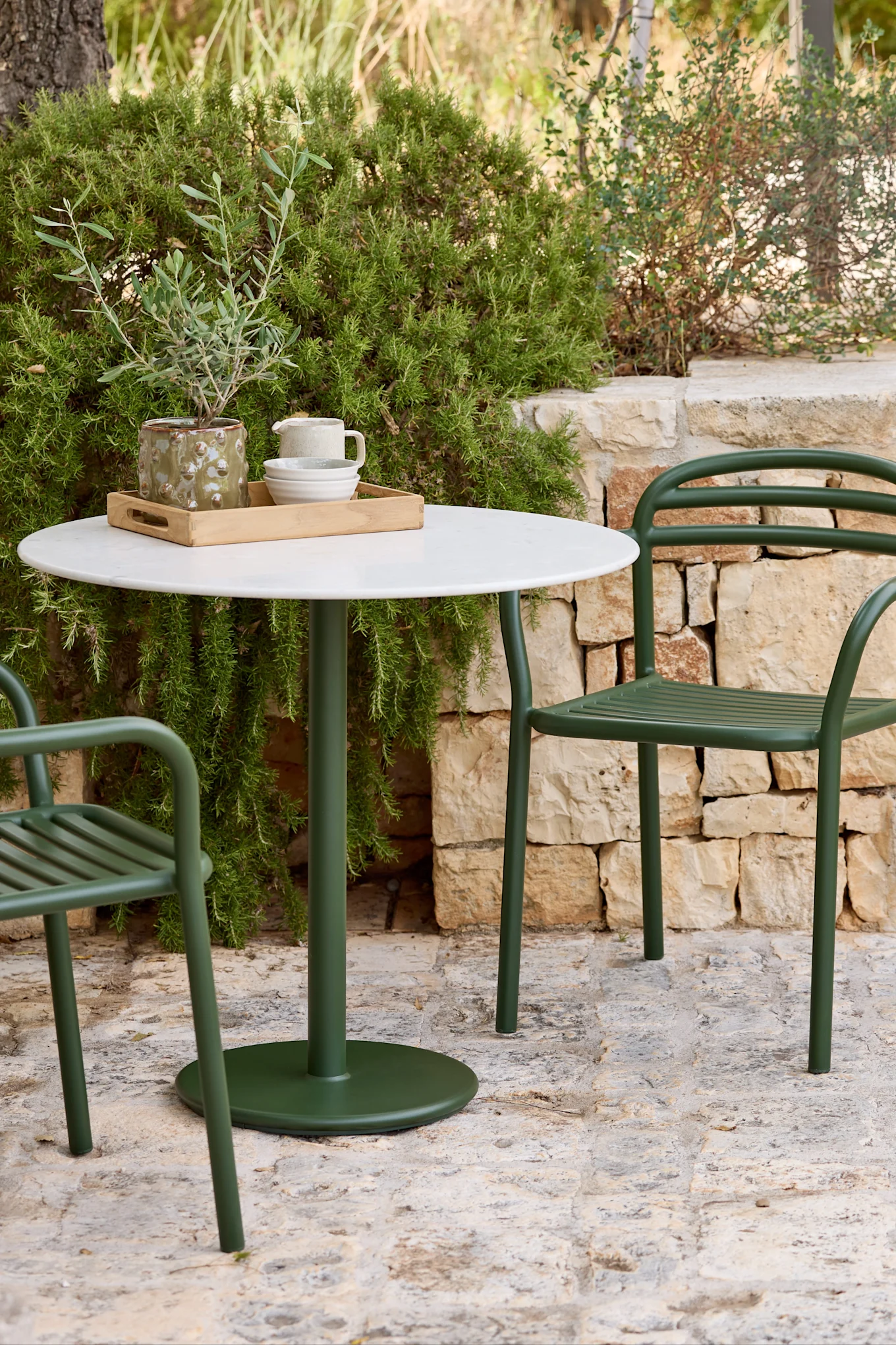 Un ensemble de salle à manger extérieur avec une table ronde blanche et deux chaises en métal vert foncé sur une terrasse en pierre, entouré d'un feuillage vert luxuriant.