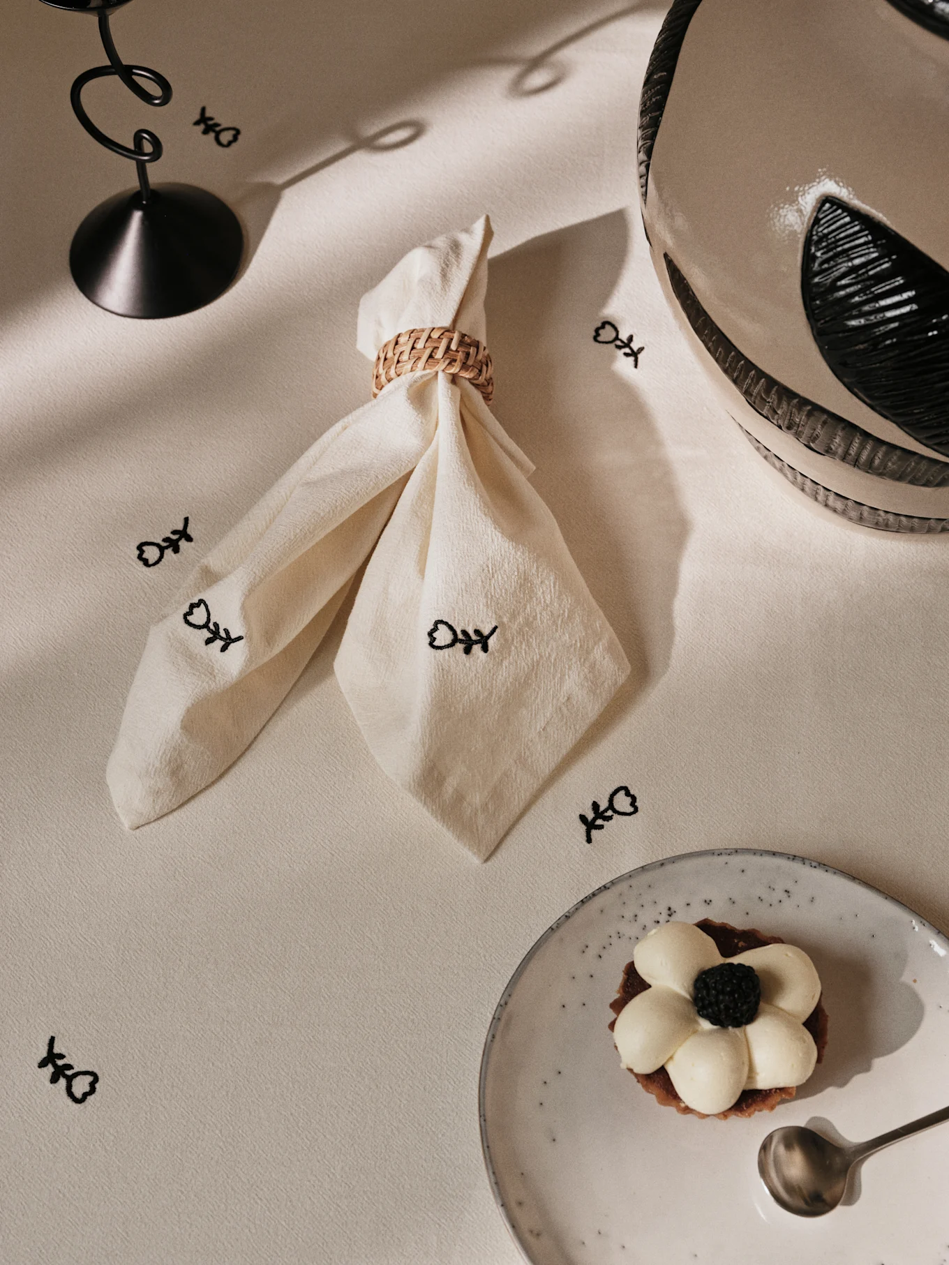 Table dressée avec une serviette crème dans un anneau tressé, des symboles d'arêtes de poisson noirs, un support métallique torsadé et une tarte aux mûres sur une assiette mouchetée.