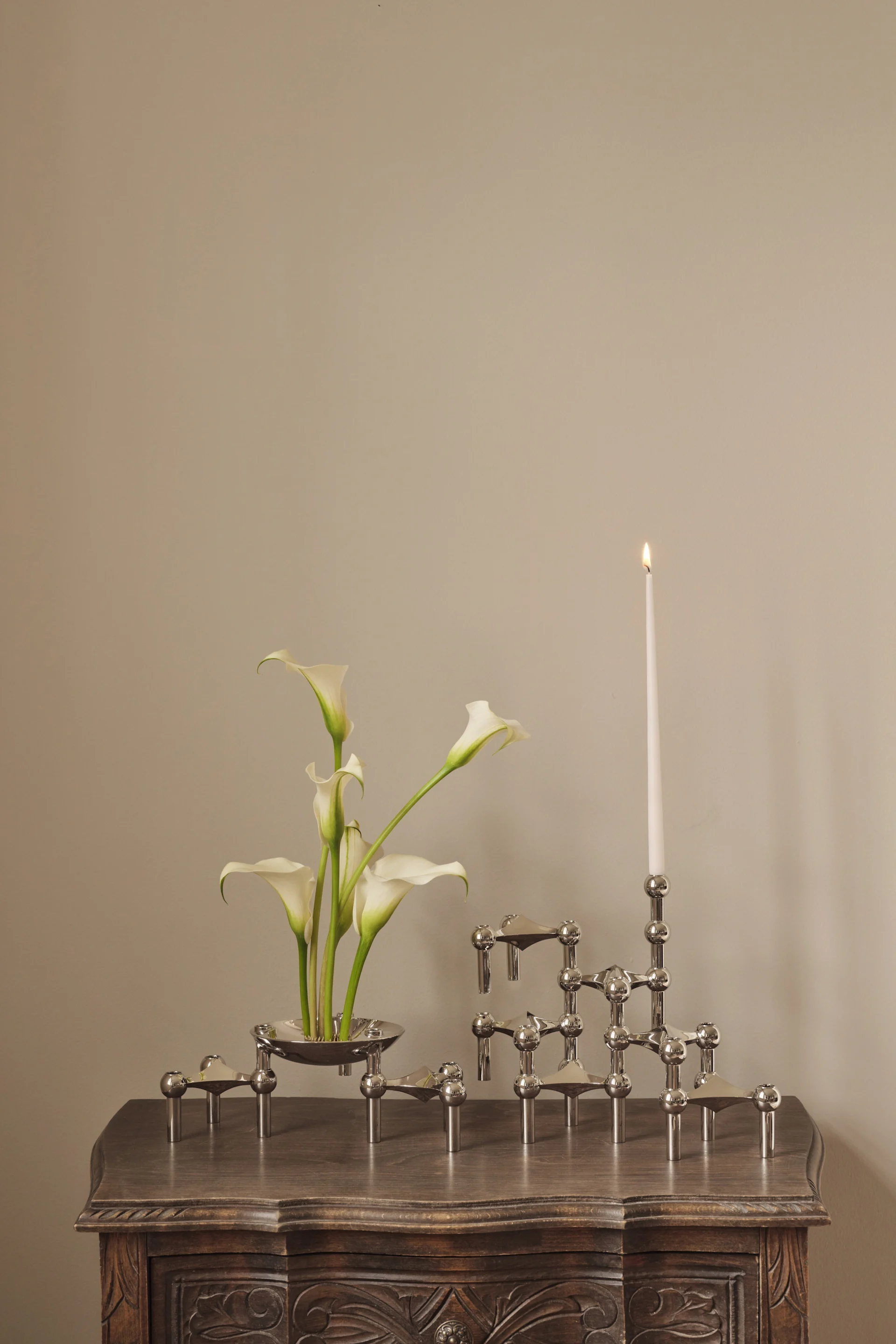 Des lis calla blancs dans un bol en argent, à côté d'un bougeoir modulaire en argent sur lequel est posée une bougie blanche allumée, le tout posé sur une table en bois sculpté.