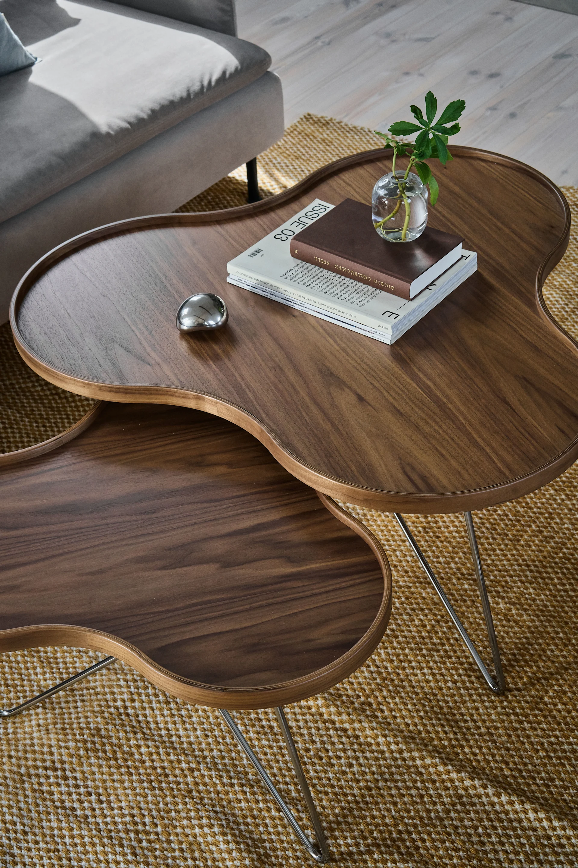 Deux tables basses gigognes en bois foncé, en forme de haricot, sur un tapis à motifs, avec une plante, des livres et un objet métallique.