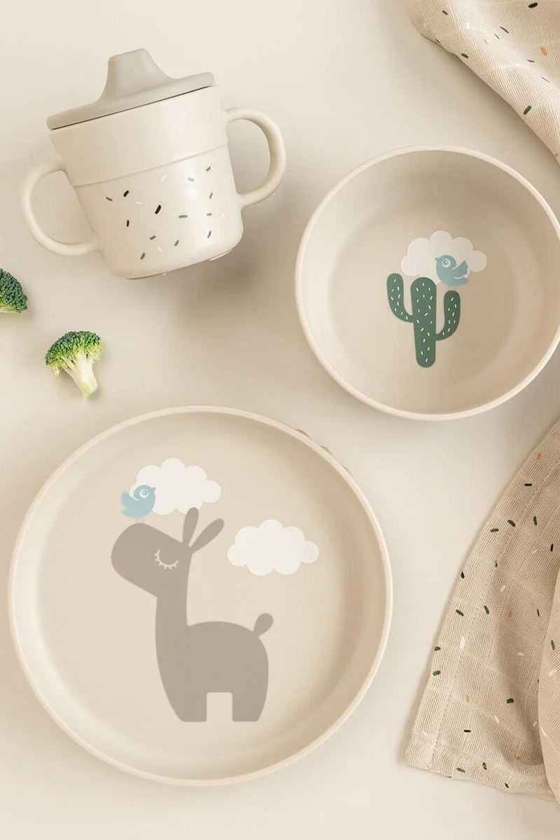 Un service de vaisselle pour enfants beige clair avec une tasse à bec, un bol avec un cactus et une assiette avec un lama et des oiseaux.