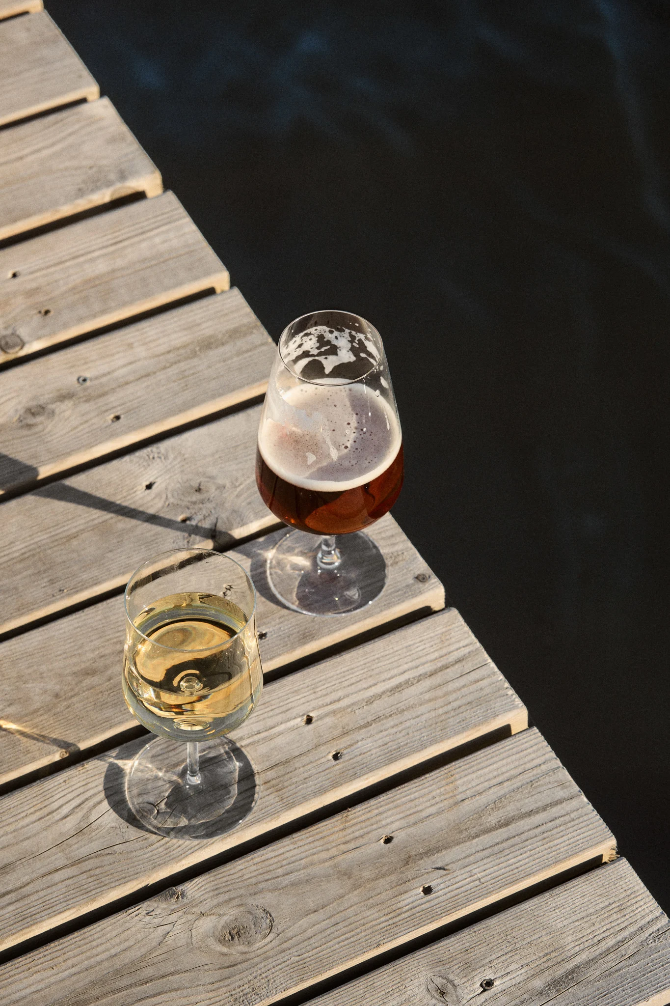 Deux verres, un de vin blanc et un de bière, sur un quai en bois près d'une eau sombre sous le soleil.