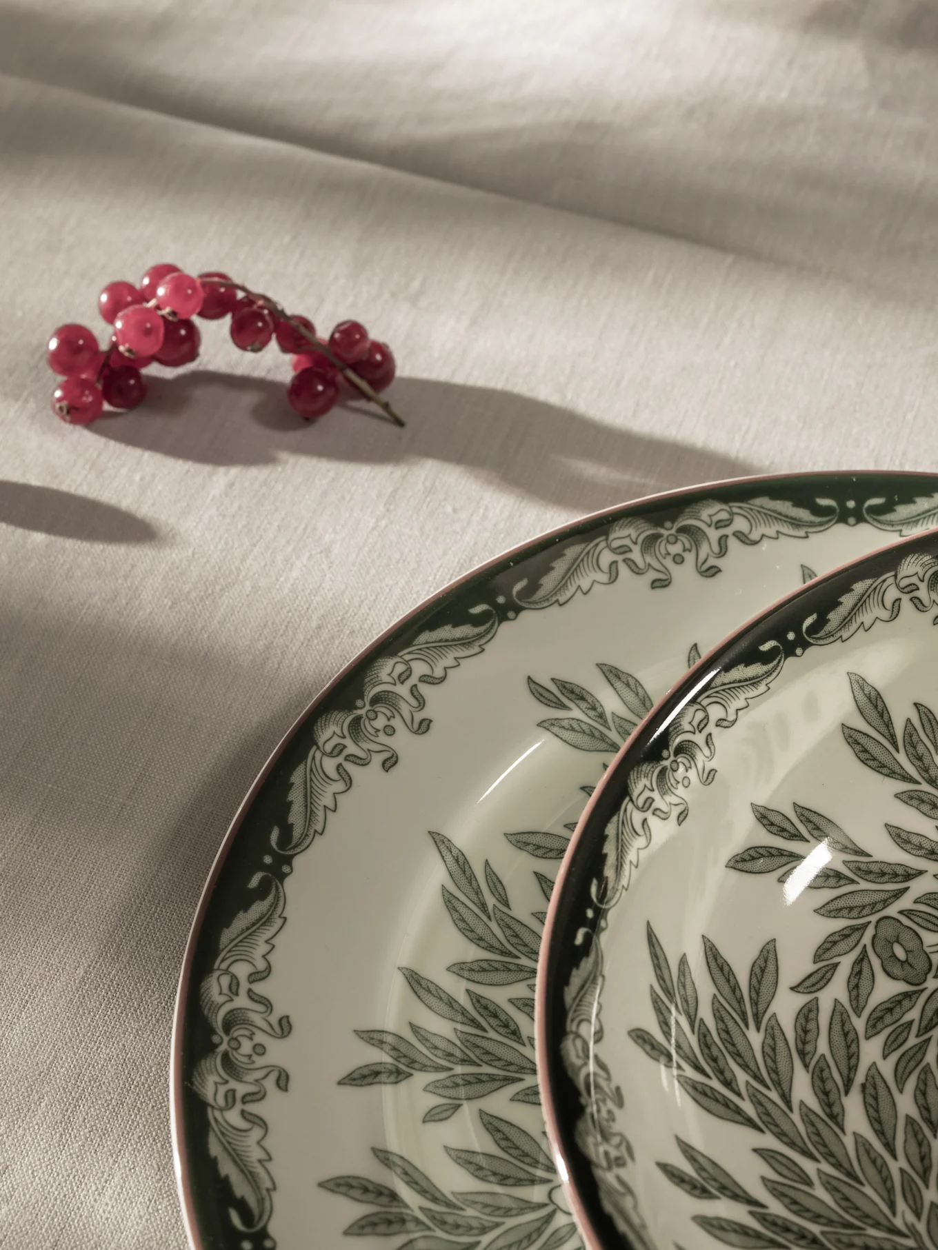 Groseilles rouges sur une nappe claire à côté de deux assiettes blanches avec des motifs de feuilles vertes et des bords roses.