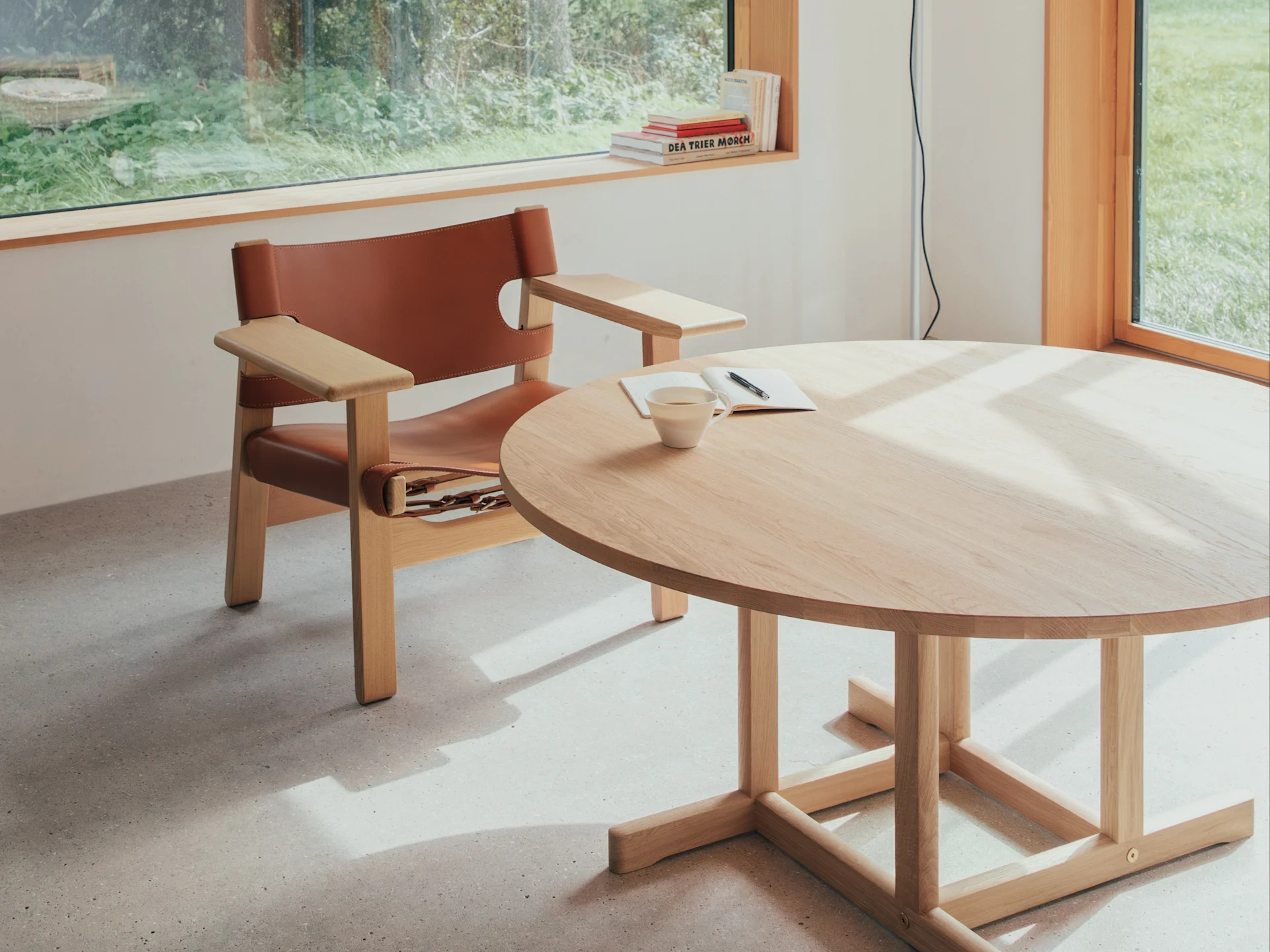Pièce lumineuse avec fauteuil en cuir marron, table ronde en bois clair avec tasse et carnet, éclairée par une fenêtre donnant sur la verdure.