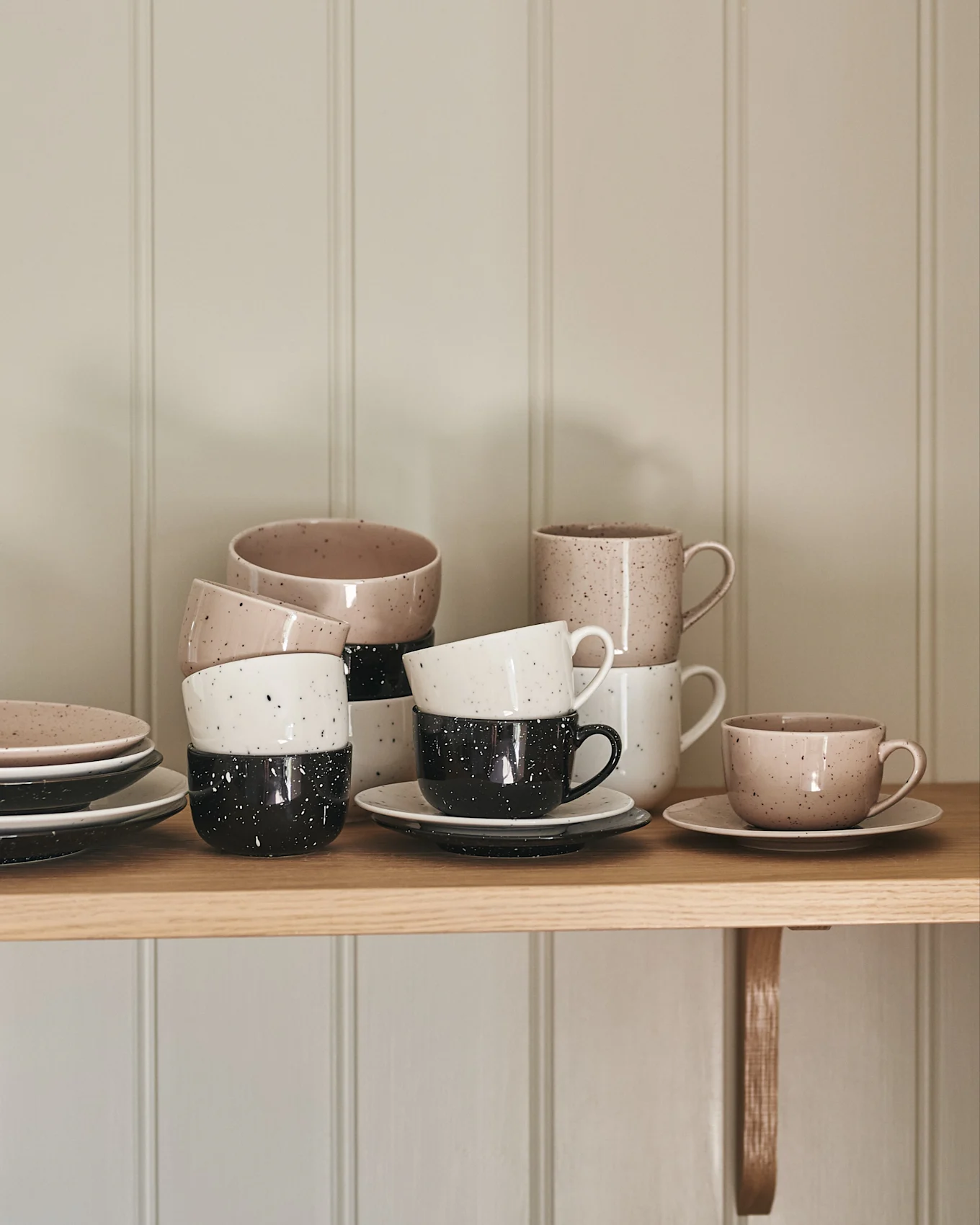 Tasses et assiettes en céramique empilées, beiges, blanches et noires mouchetées sur une étagère en bois.
