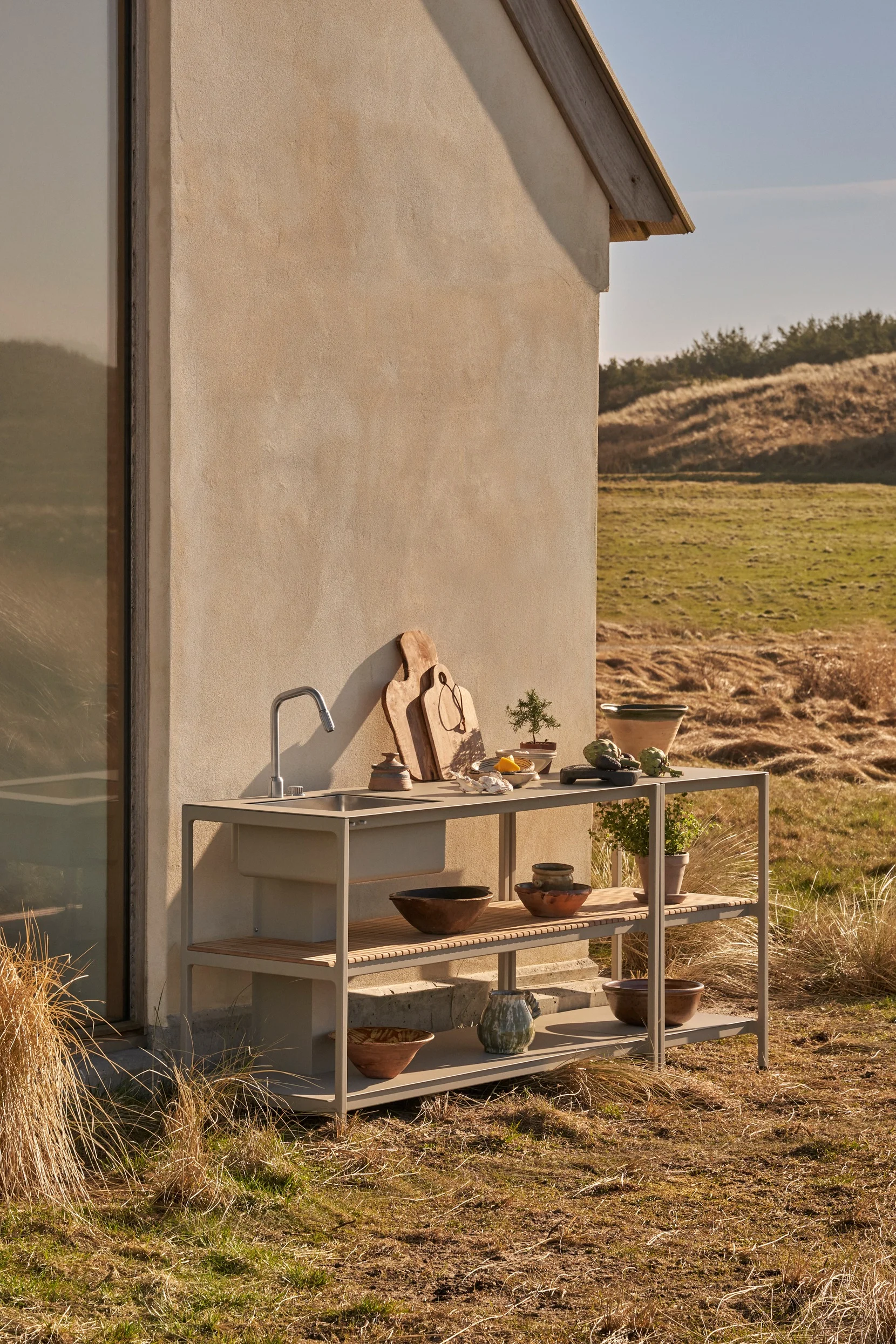 Unité de cuisine extérieure avec évier et étagères, décorée de planches à découper, de plantes et de bols, devant un bâtiment dans un paysage naturel.