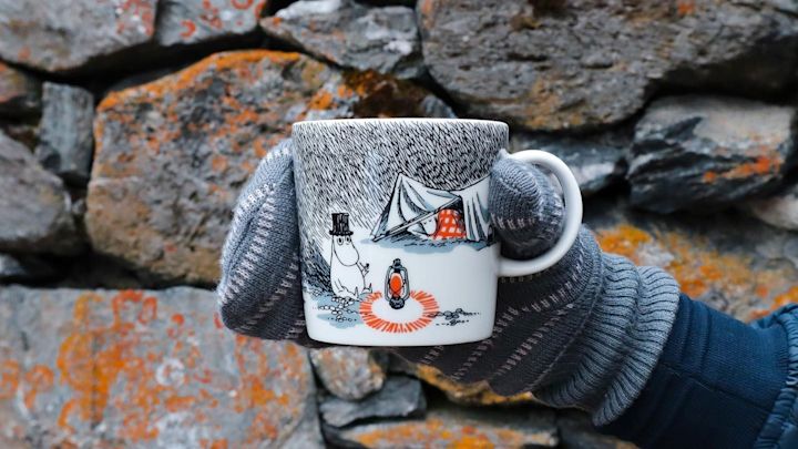 @moominbyingri tient la coupe Schlaf gut Moomin devant un mur de pierre en portant un gant.