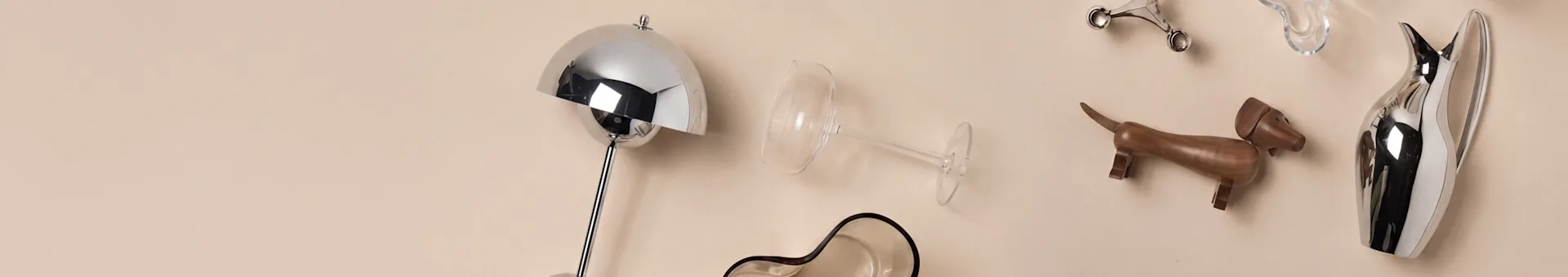 Assortiment d'objets de décoration intérieure incluant une lampe argentée, un verre à vin, un chien en bois et une carafe argentée sur un fond beige.