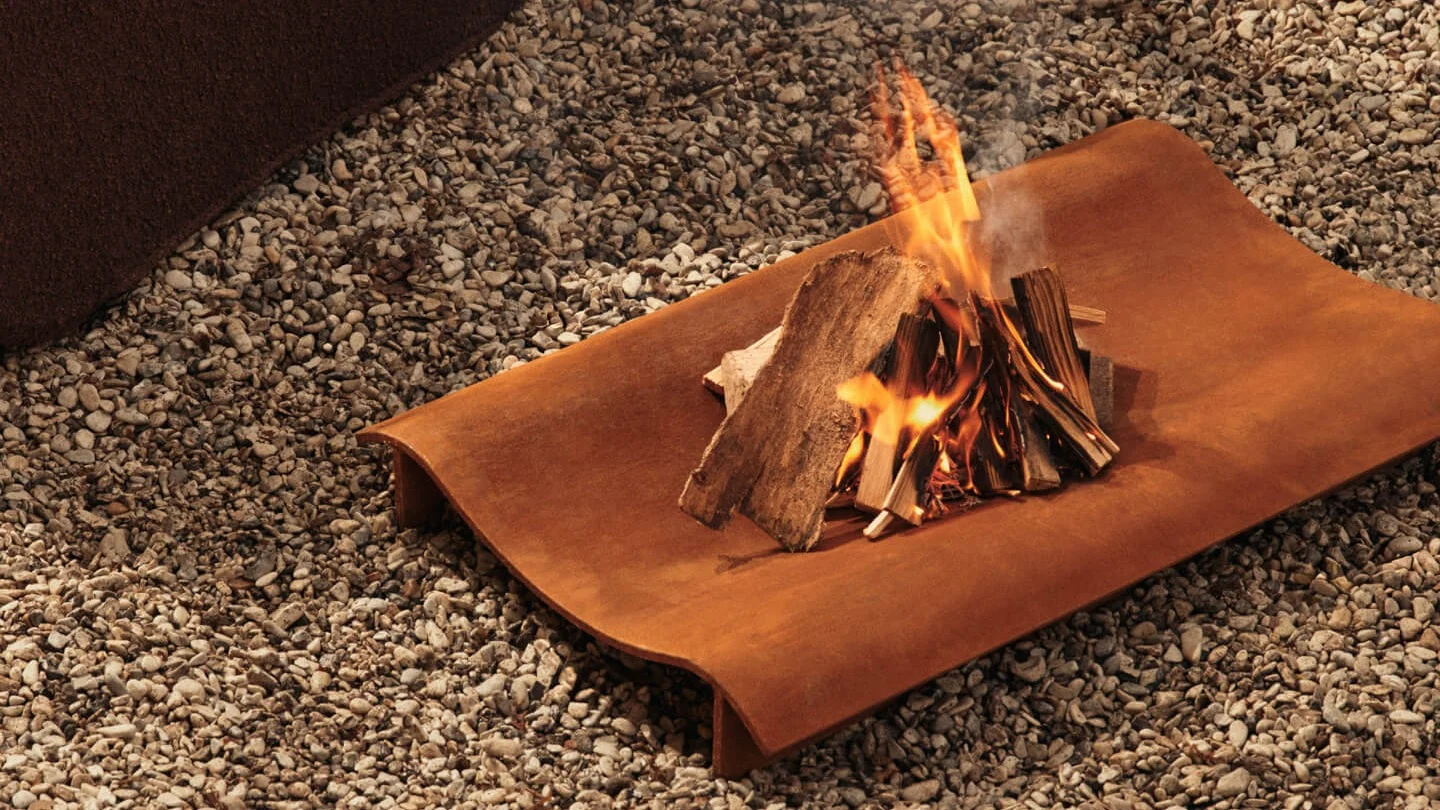 Petit feu de joie avec du bois qui brûle sur un plateau rectangulaire en métal rouillé sur du gravier.