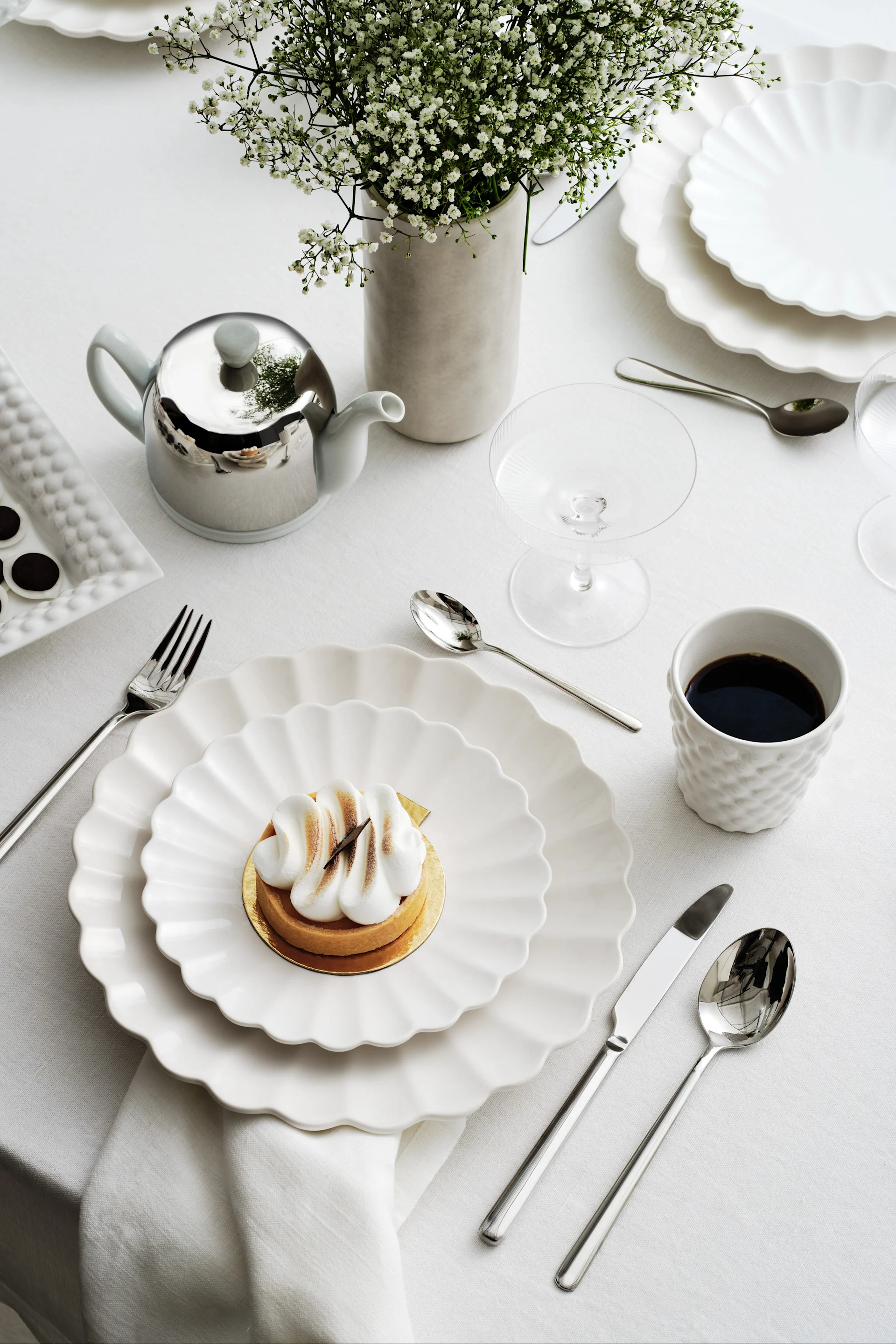 Table élégante avec assiettes festonnées blanches, une tarte meringuée, du café, une théière argentée et des gypsophiles.
