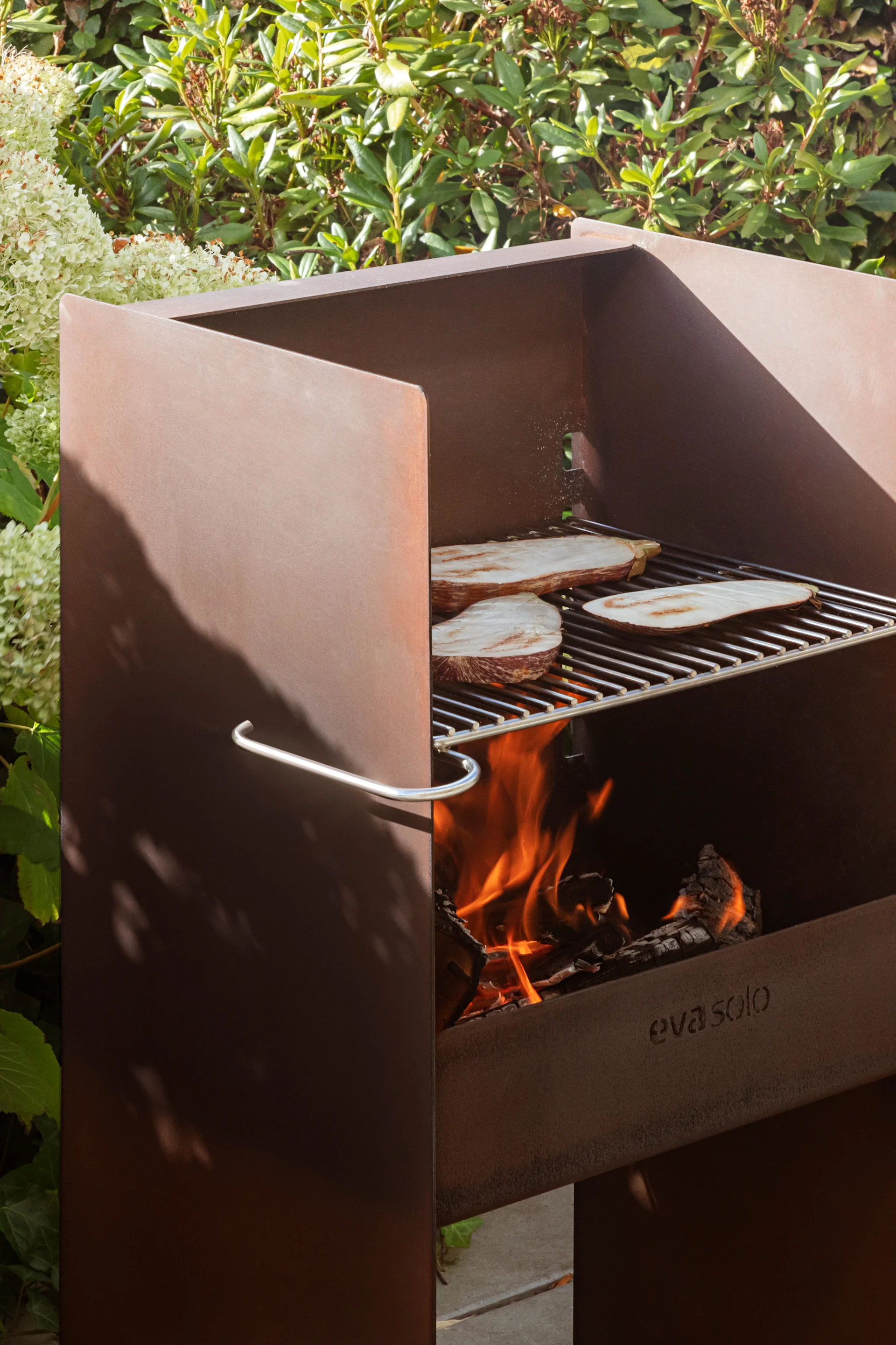 Barbecue d'extérieur en acier Corten avec des tranches d'aubergine grillées sur une grille au-dessus d'un feu ouvert, entouré de plantes vertes.