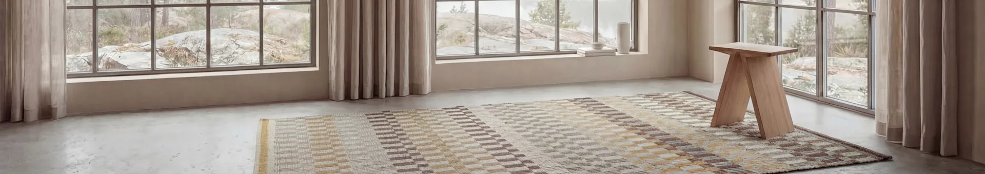 Une pièce avec de grandes fenêtres dévoilant un paysage rocheux. Un tapis à motifs et un tabouret en bois clair sur un sol gris, avec de la décoration sur le rebord.