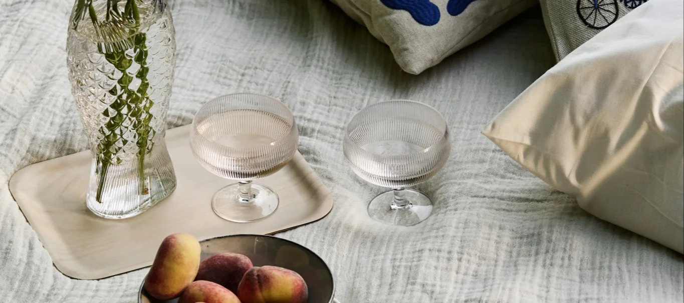 Une mise en place de pique-nique avec un vase en forme de poisson, deux verres à pied nervurés et des pêches posées sur une couverture claire.