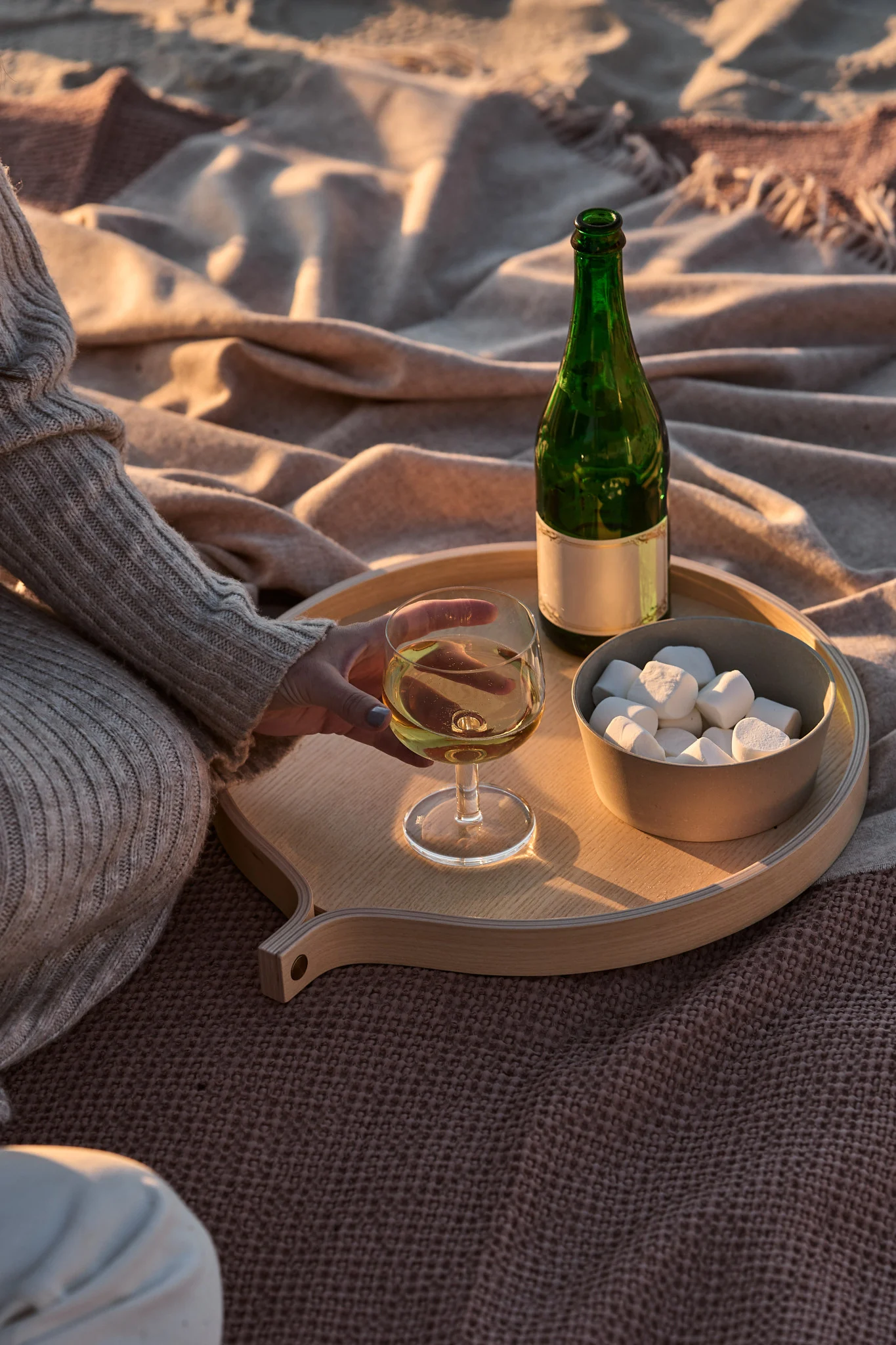 Une personne tient un verre de vin blanc sur un plateau en bois, à côté d'une bouteille verte et de guimauves disposées sur des couvertures, pendant l'heure dorée.