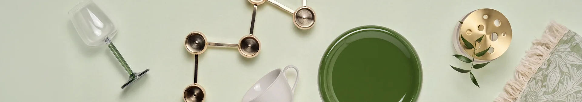 Vue de dessus: verre à pied vert, décor sculptural doré, tasse blanche, assiette verte, objet en laiton avec feuilles, textile vert frangé.