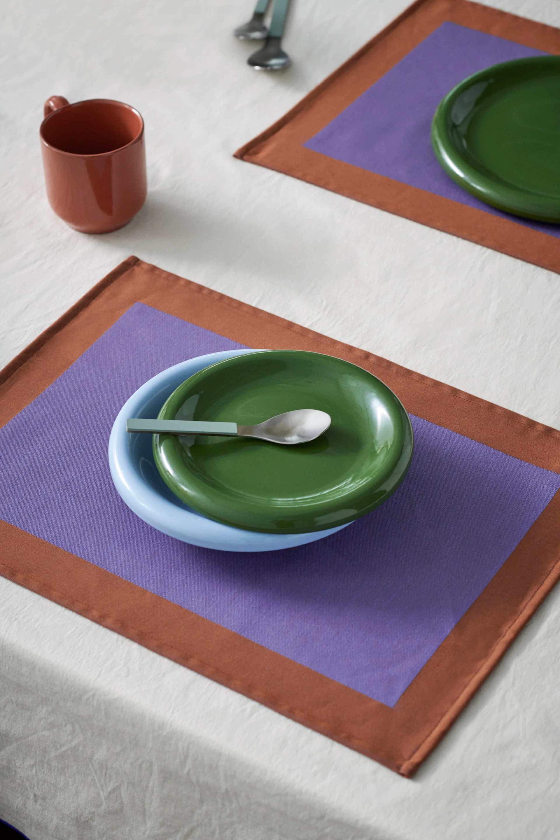 Table dressée: bol bleu, assiette verte, cuillère sur set de table violet et marron. Tasse marron sur nappe blanche.