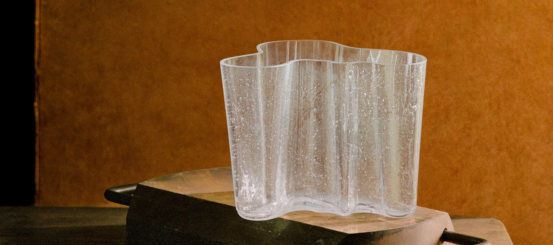 Un vase en verre clair et ondulé avec des mouchetures texturées sur une surface rustique sombre devant un fond marron chaud.