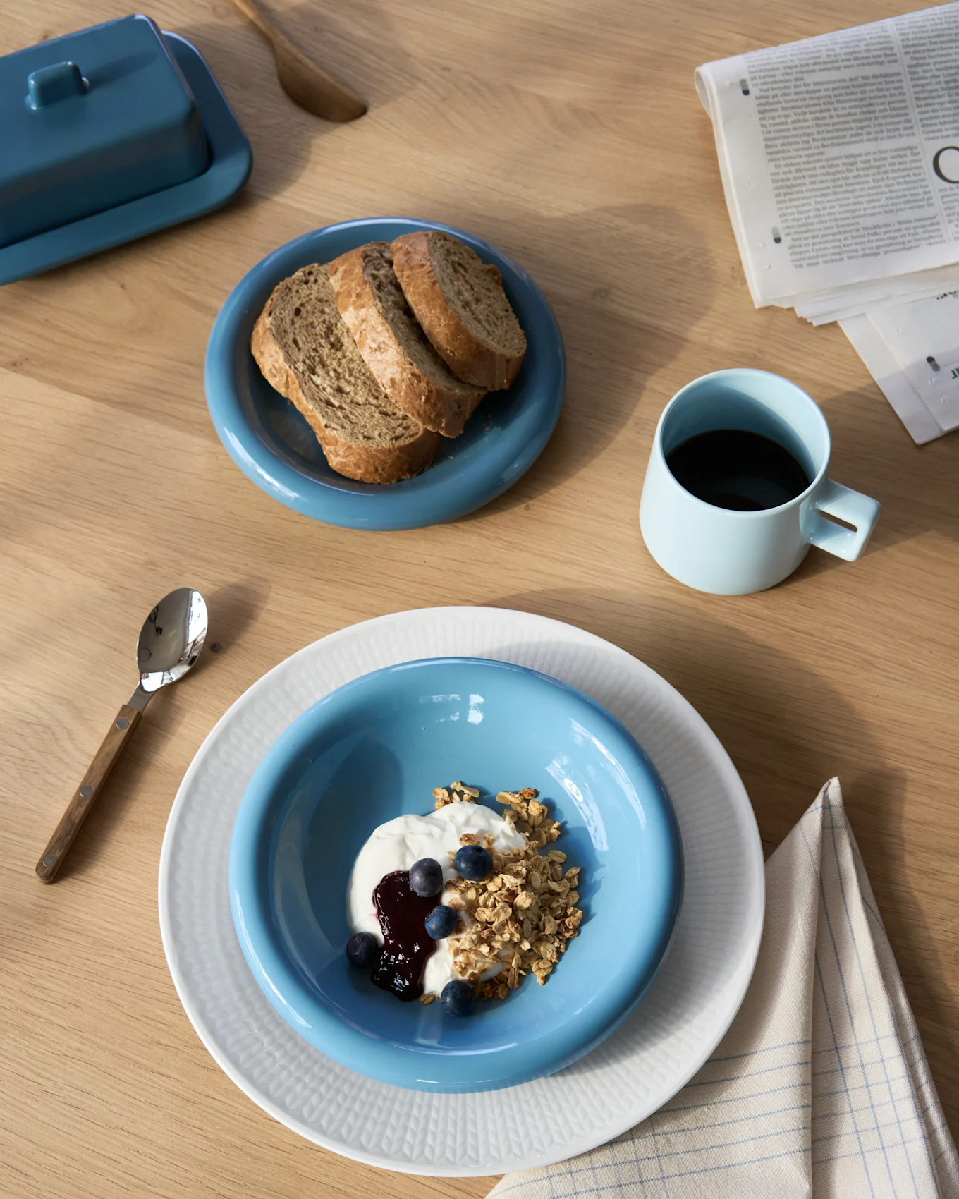 Vue plongeante d'un petit-déjeuner avec yaourt et granola, pain noir, café et beurrier sur une table en bois.