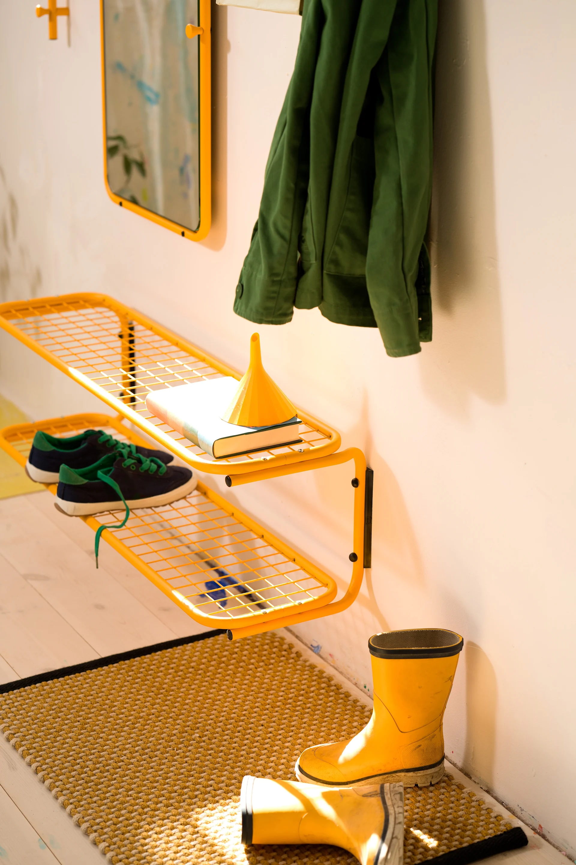 Entrée avec étagère à grille jaune, veste verte, baskets bleues et bottes en caoutchouc jaunes sur un tapis texturé.
