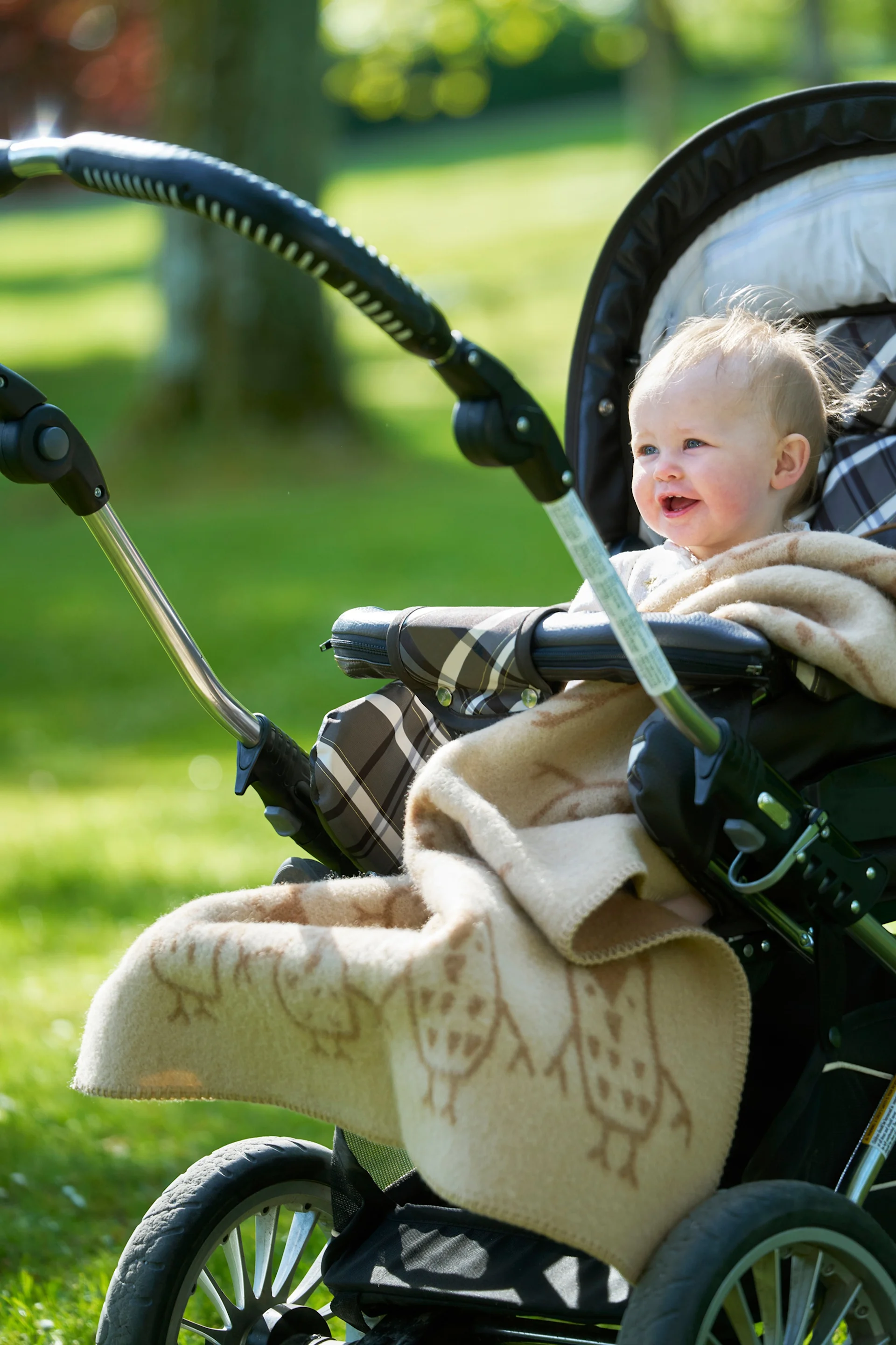 Un bébé souriant aux cheveux clairs, enveloppé dans une couverture à motif de hiboux, est assis dans une poussette en plein air dans un parc ensoleillé.