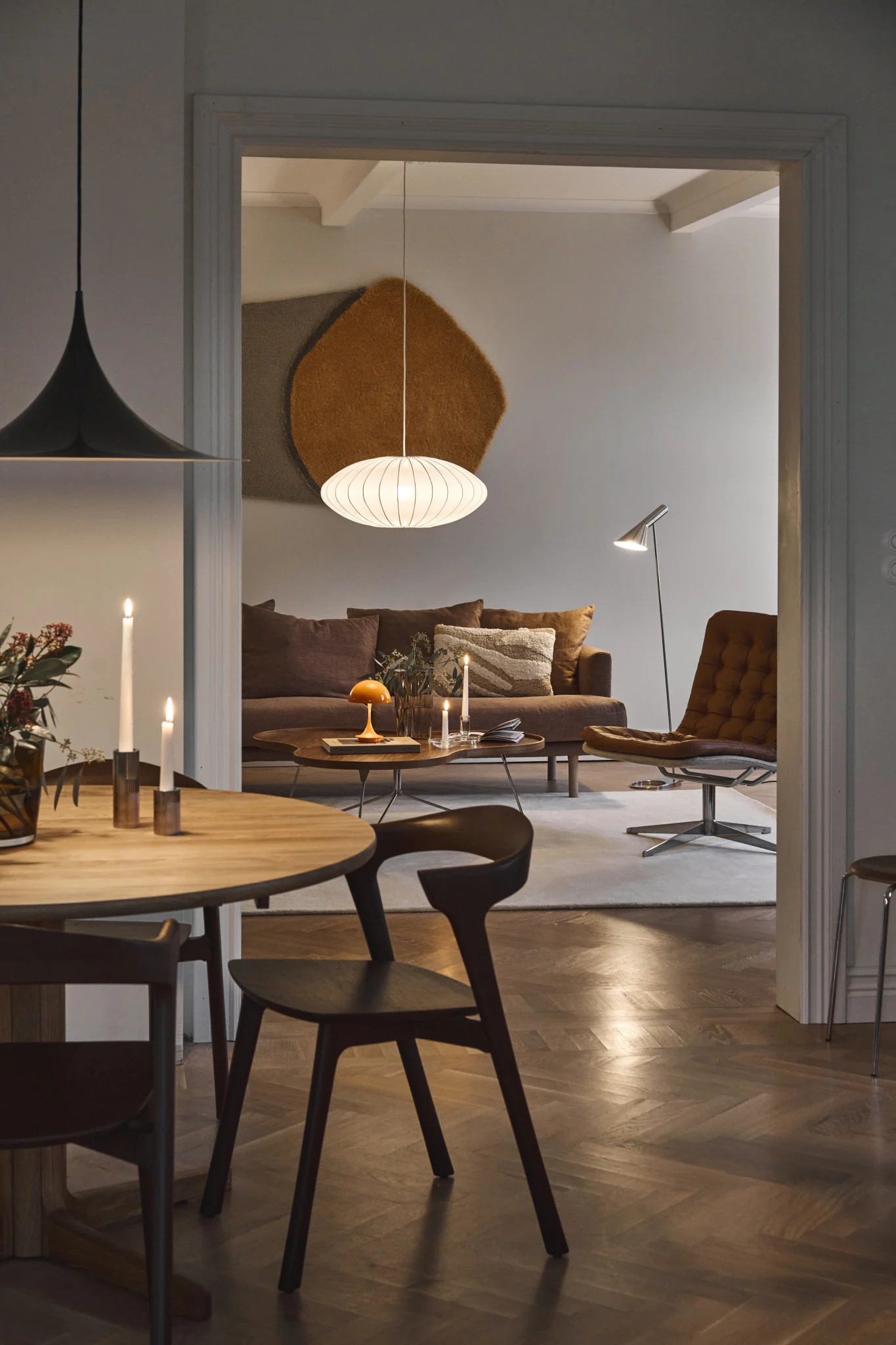Vue d'une salle à manger vers un salon avec un canapé marron, un fauteuil en cuir et diverses lampes, dont une suspension ovale blanche.