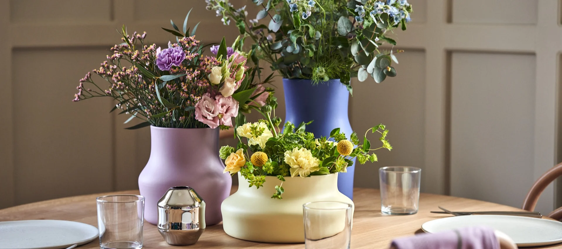 Sur une table dressée, trois vases colorés de la collection Dorotea de Gense sont garnis de fleurs coupées multicolores.