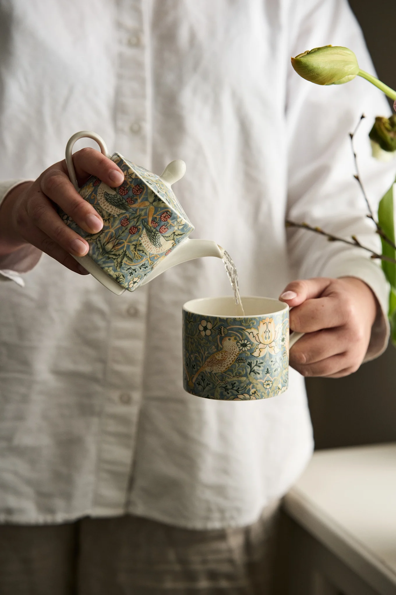 Mains versant d'une théière bleue à motif d'oiseaux et de baies dans une tasse assortie.