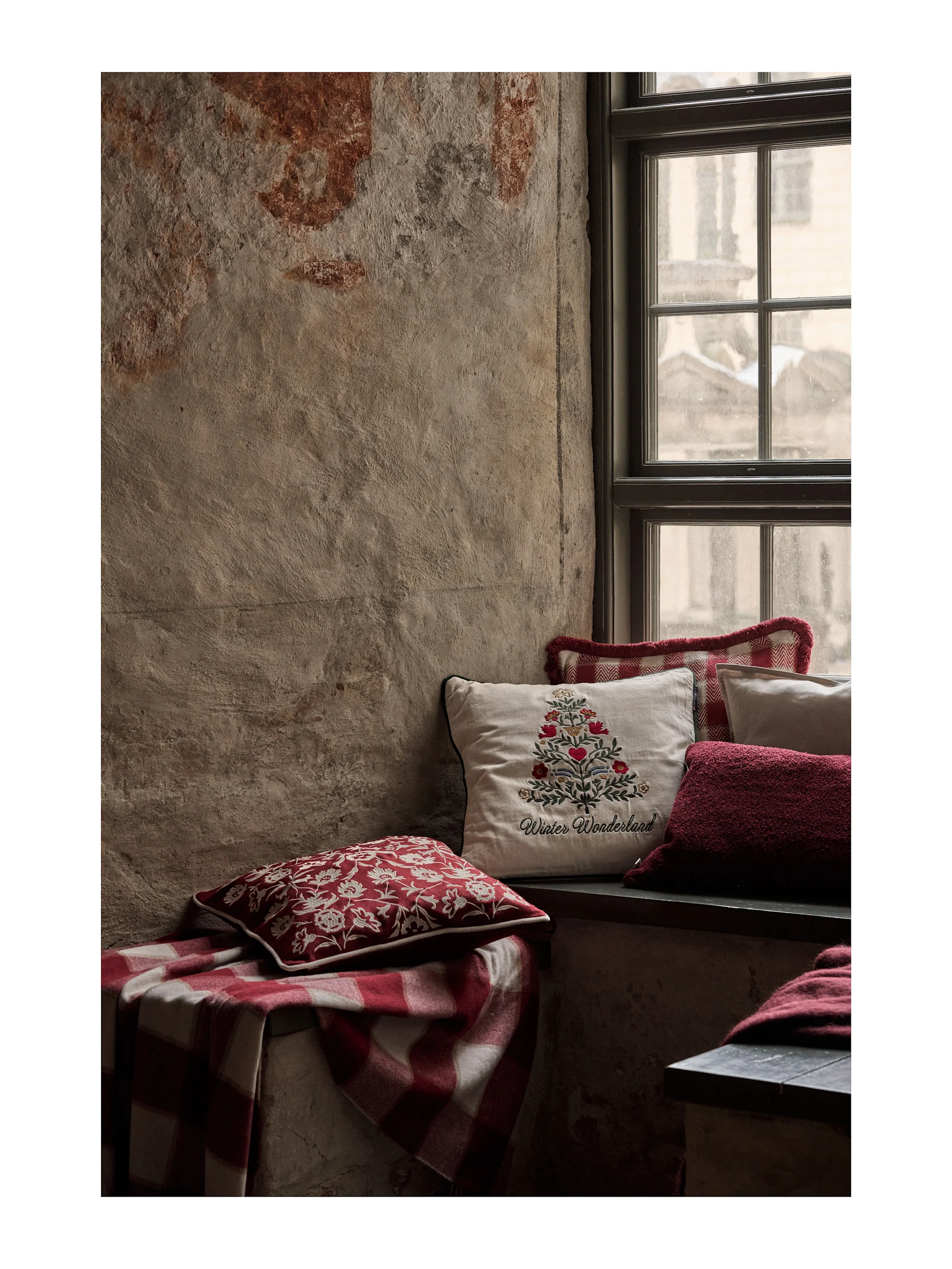 Coin de fenêtre douillet avec des coussins festifs rouges et blancs et une couverture à carreaux. Un coussin crème brodé 'Winter Wonderland'.