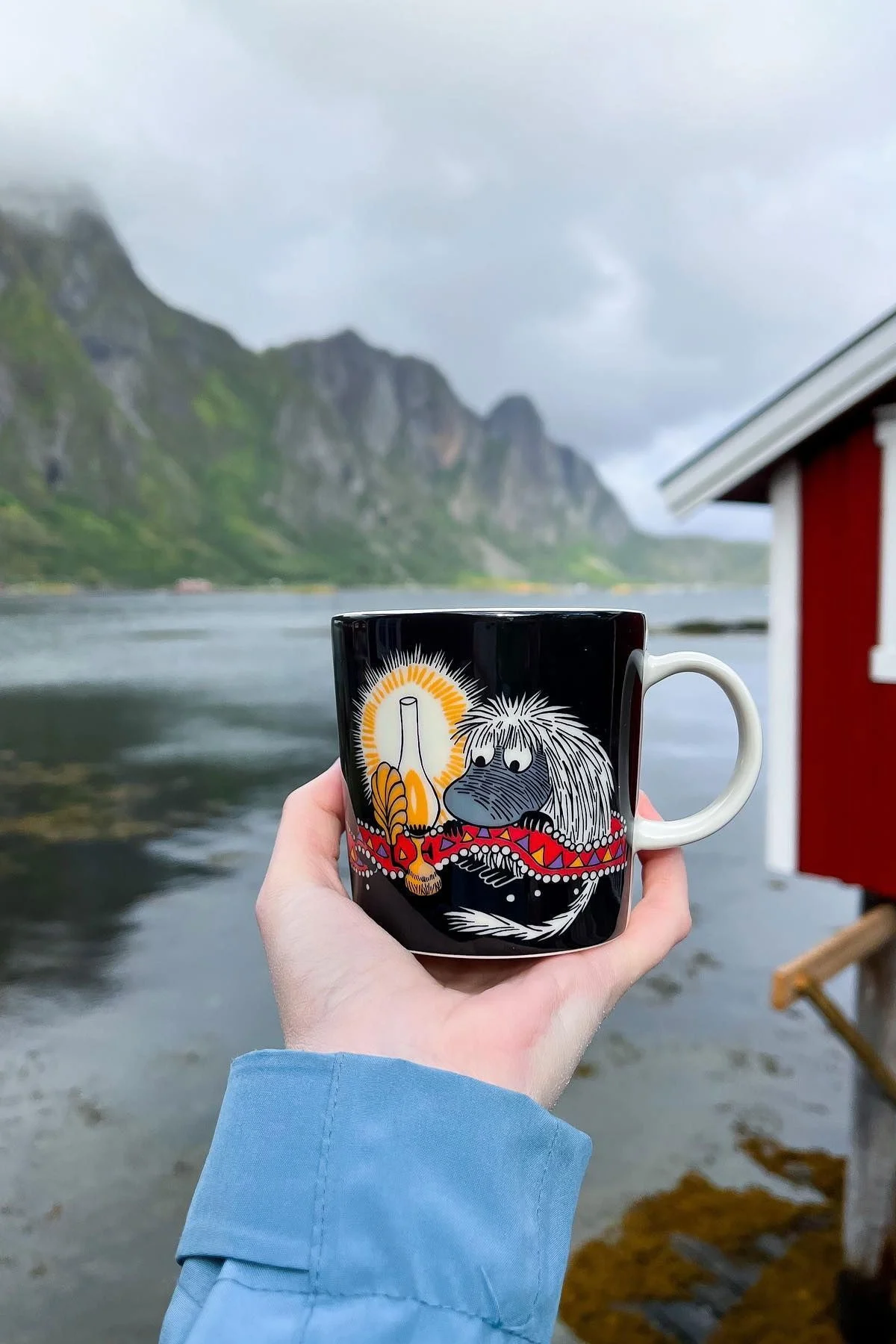 @moominbyingri tient la tasse de moomin Urahn dans sa main, avec un paysage norvégien accidenté en arrière-plan.