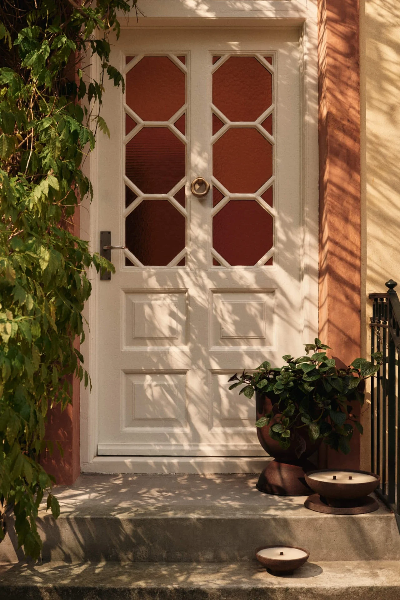 Porte d'entrée blanche avec panneaux de verre octogonaux, vignes vertes à gauche et jardinière avec deux bougies sur les marches.