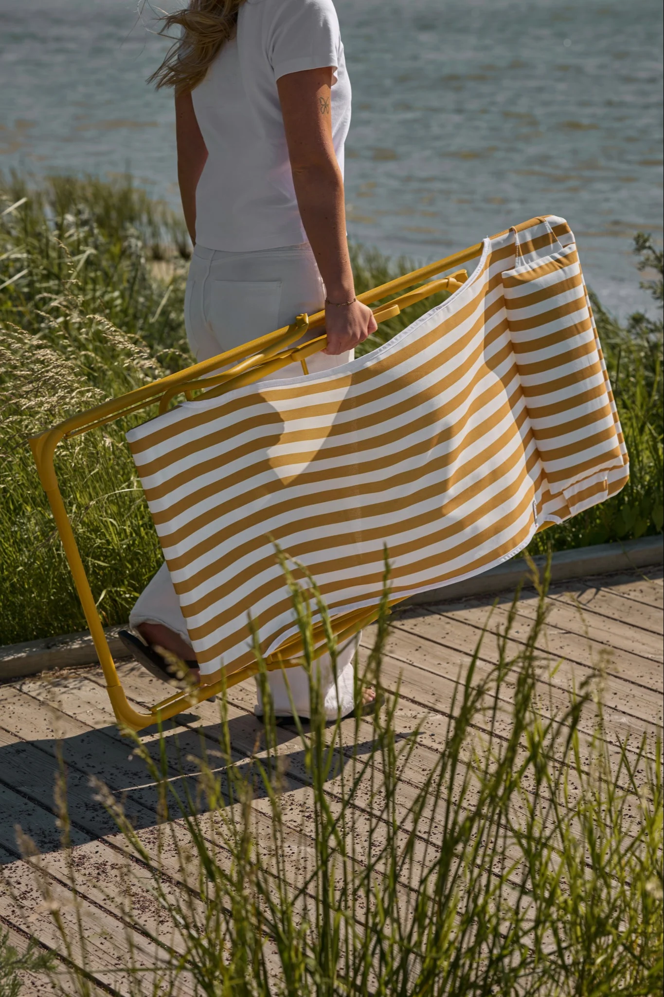 Personne en blanc porte une chaise de plage pliable rayée jaune et blanc sur une promenade en bois avec de hautes herbes au bord de l'eau.
